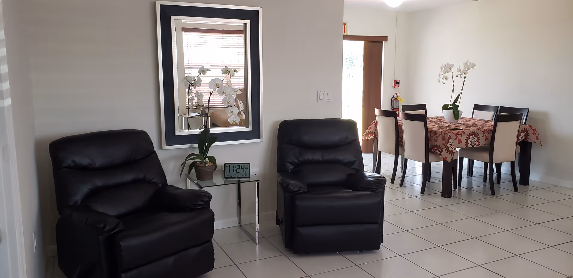 A living area with two black leather recliners separated by a small glass side table holding a potted orchid and a digital clock. A large mirror hangs on the wall above the table. In the background, there is a dining area with a table covered by a red patterned tablecloth and six chairs. Another potted orchid is placed on the dining table. The floor is tiled, and there is a door with a window letting in natural light.