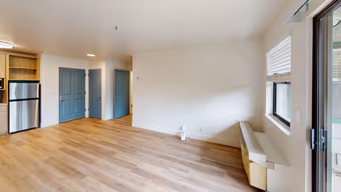 Empty living area of a senior apartment with wood-look flooring, blue doors, a small kitchenette with a stainless refrigerator on the left, and windows/sliding door on the right.