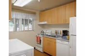 A compact kitchen with light wood cabinets, a white stove with a kettle on it, a white refrigerator, and a white washing machine. There is a window with white blinds and a sheer curtain, and various kitchen utensils and items are placed on the countertop.