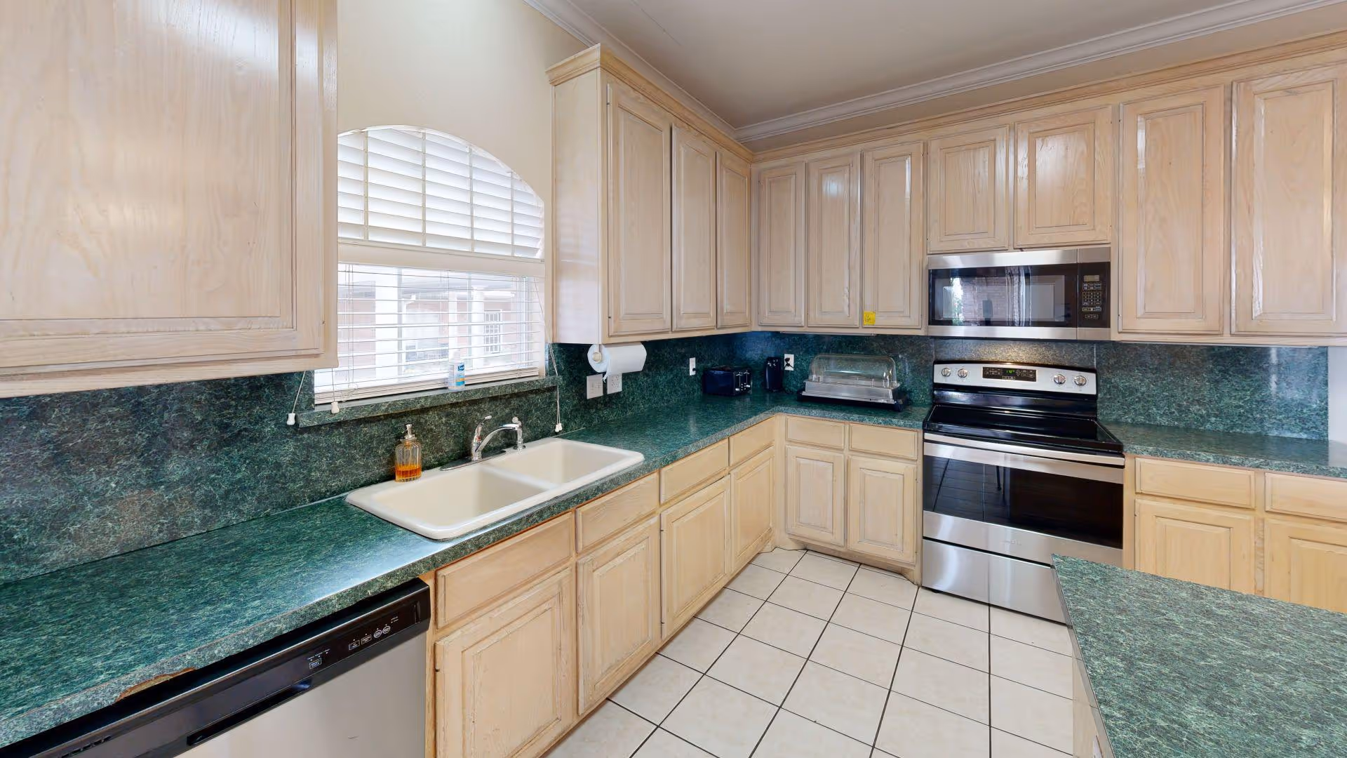 Bright kitchen with green countertops, light wood cabinets, a double sink under a window, and stainless steel oven and microwave.