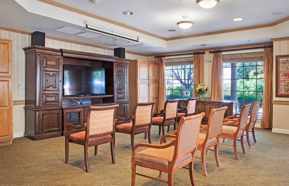A well-lit common room with a large wooden entertainment center holding a flat-screen TV. In front of the TV, there are two rows of wooden chairs with cushioned seats and backs arranged facing the screen. Large windows with brown curtains allow natural light to fill the room, and a vase with flowers sits on a table near the windows.