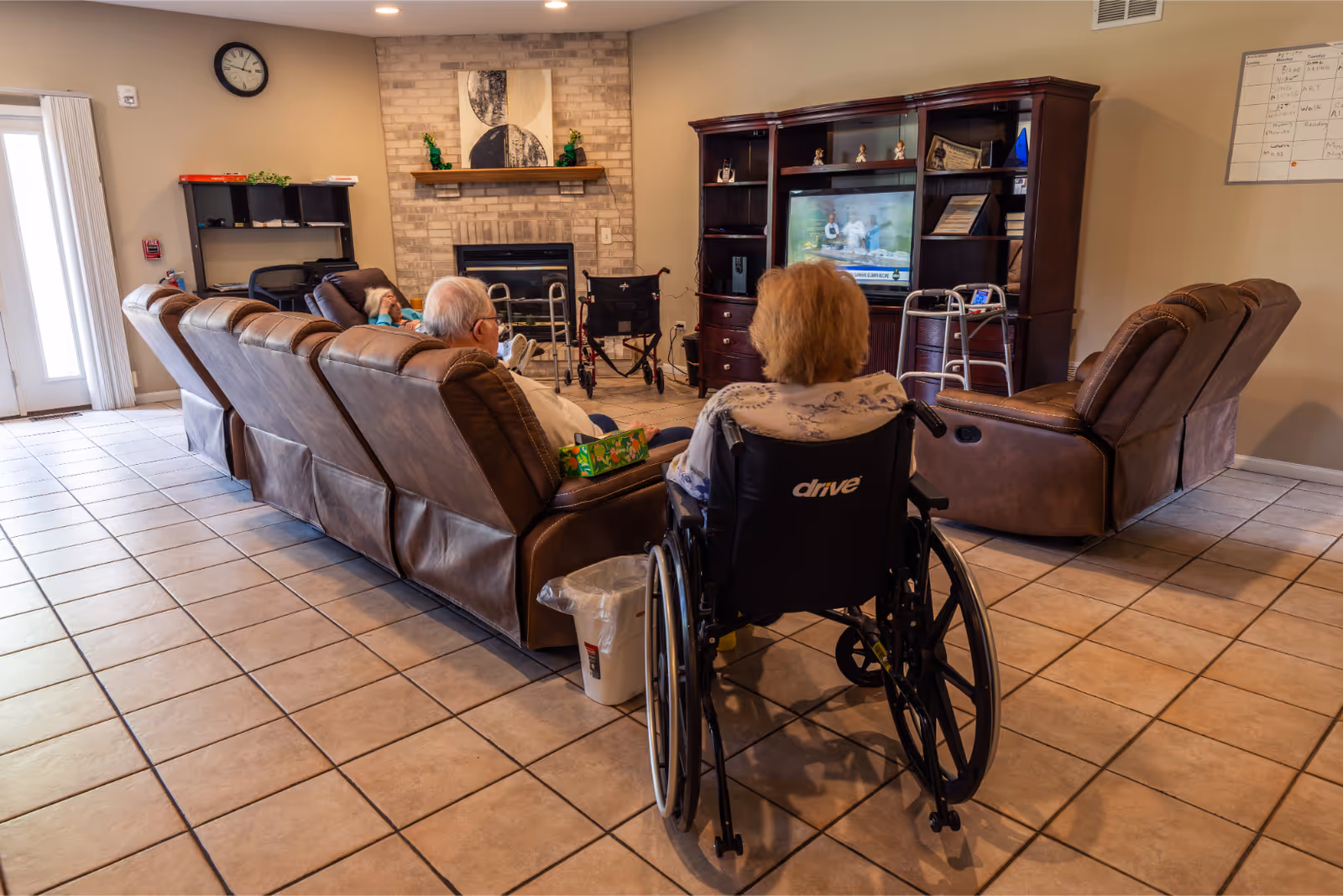 A senior living facility common area with several elderly individuals seated and watching television. The room features a row of brown recliner chairs, a person in a wheelchair, a fireplace with a mantel, and a wooden entertainment center with a TV. The floor is tiled, and there is a clock on the wall.