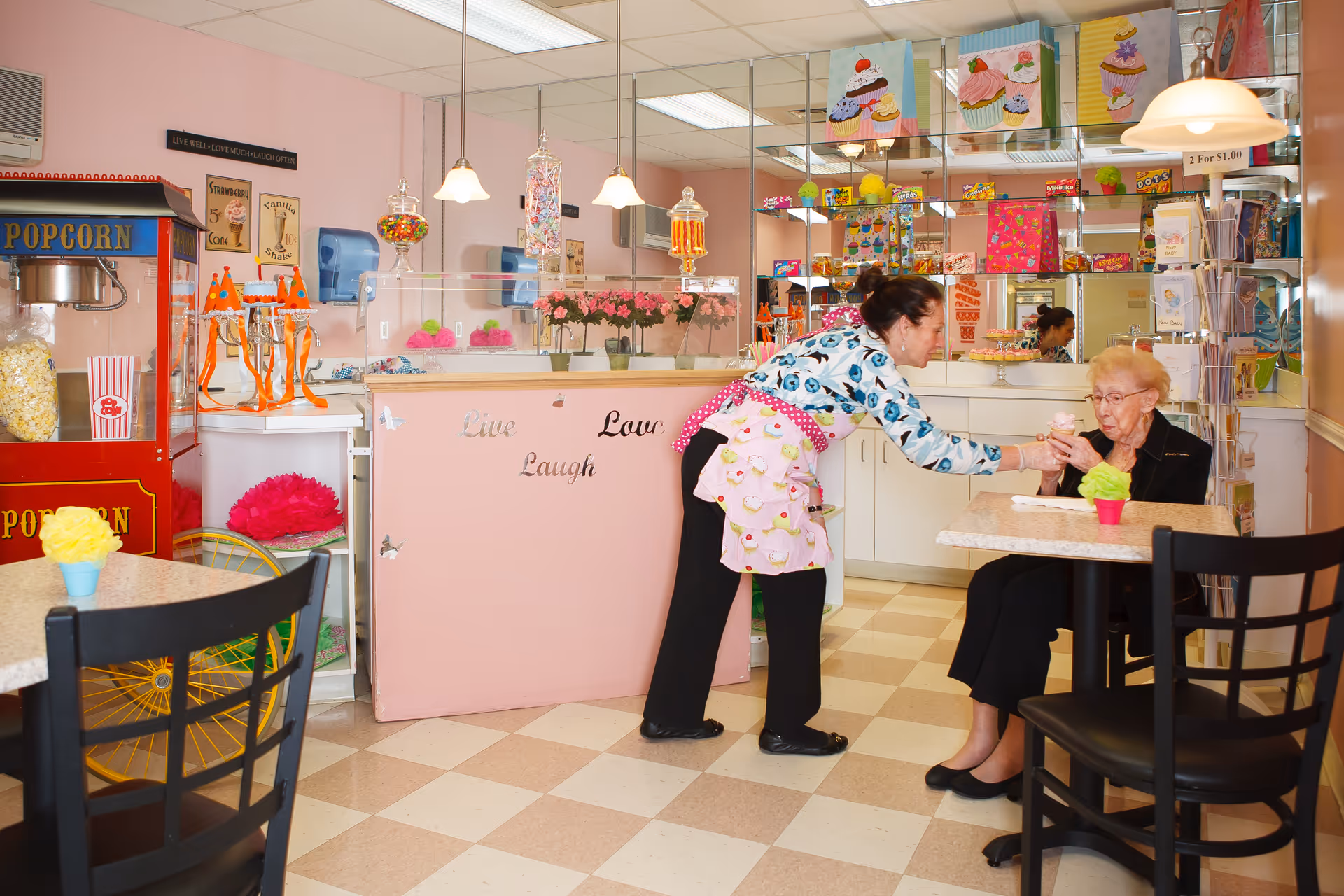 A staff member hands a dessert to an elderly woman in a pink-themed snack shop/cafe inside a senior living facility.