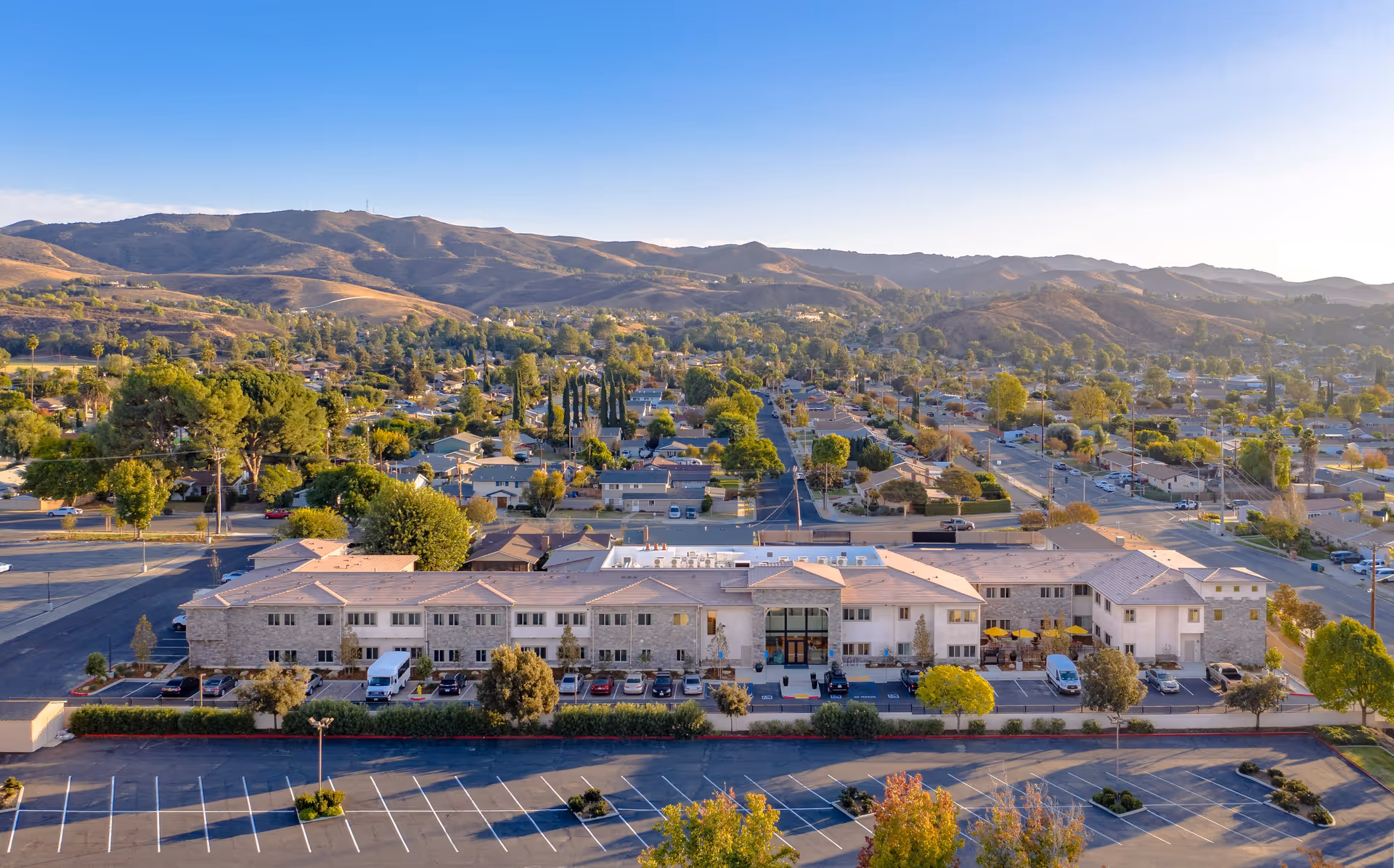 Aerial view of a long two-story senior living building with a parking lot in front and a suburban neighborhood and hills beyond.