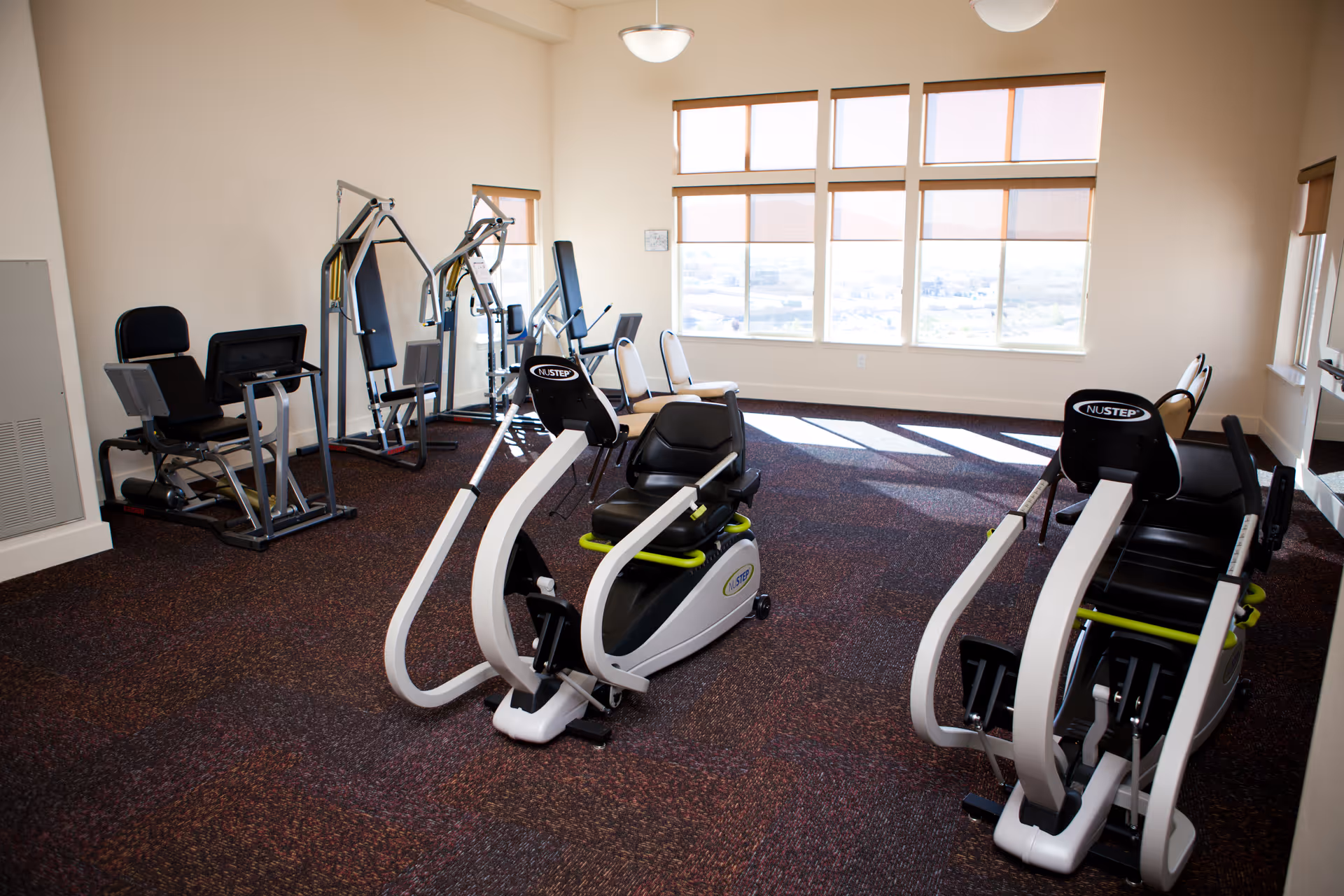 Bright fitness room with several exercise machines and large windows letting in natural light.
