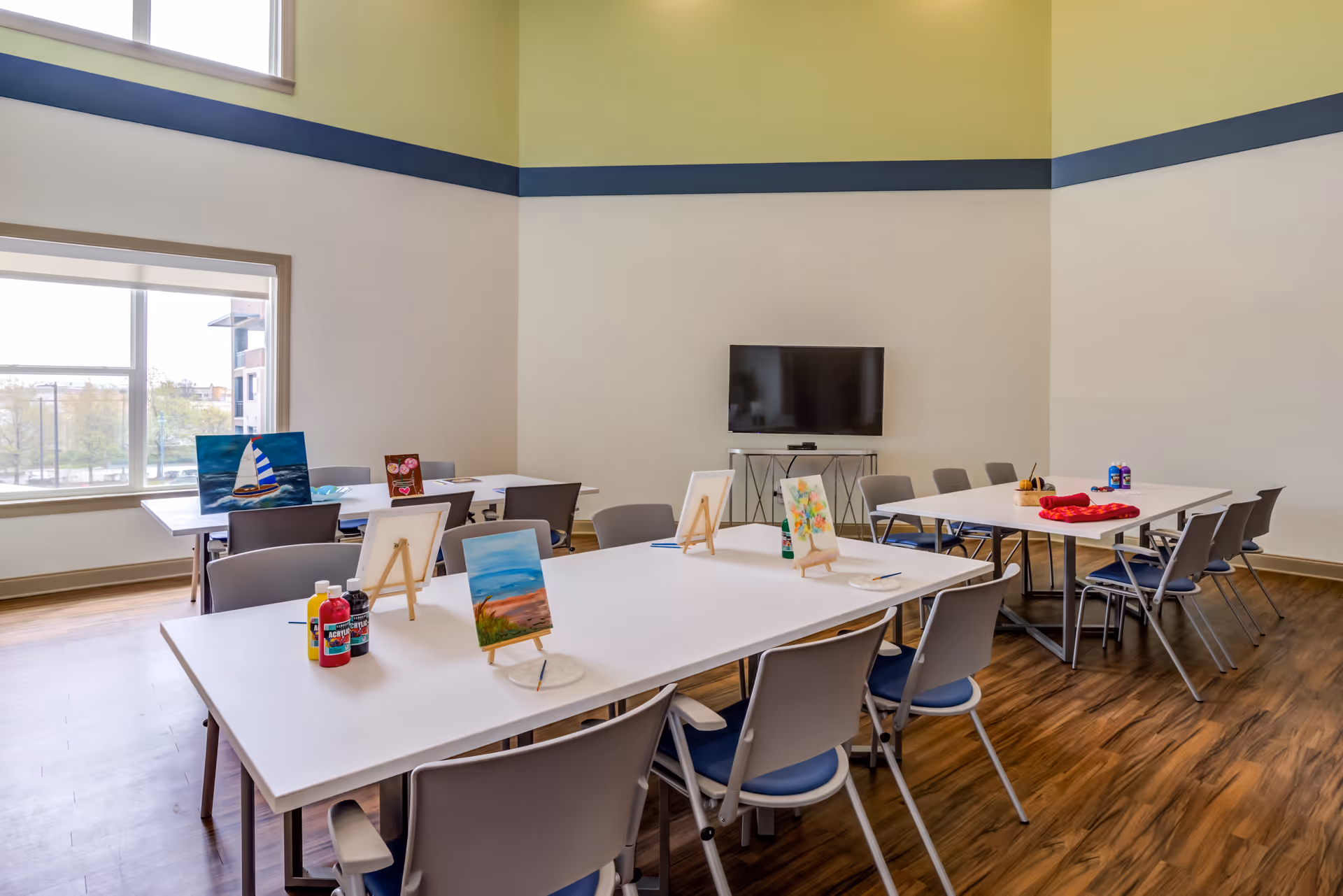 A bright activity room with high ceilings and large windows. Several white tables are arranged with chairs around them. On the tables are small easels holding paintings, paint bottles, and brushes, suggesting an art class setup. A flat-screen TV is mounted on the wall in the background.