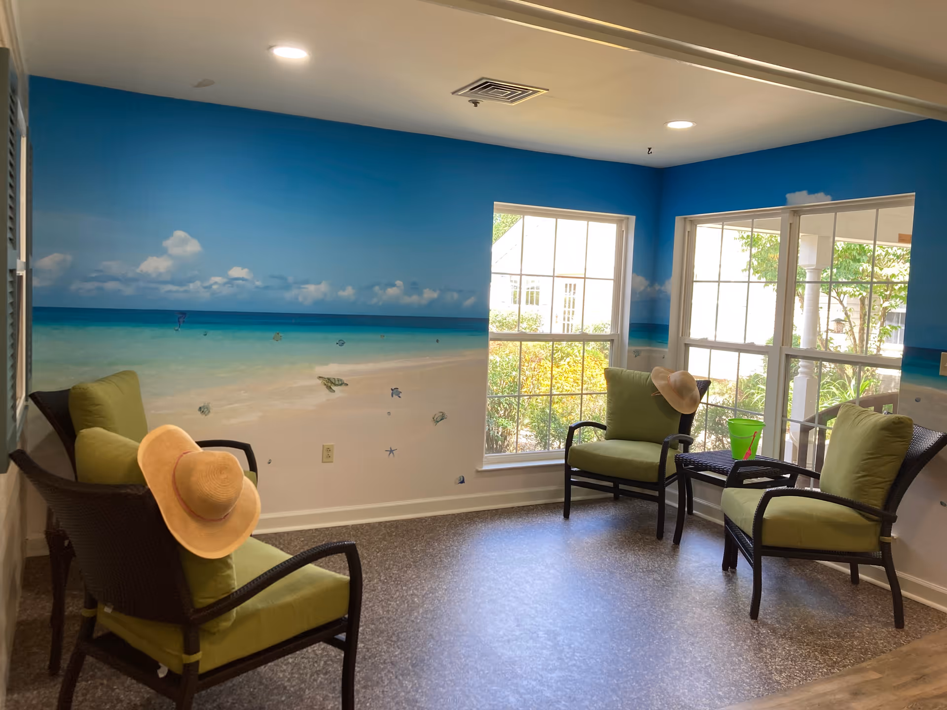 A cozy seating area with four wicker chairs featuring green cushions, two of which have straw hats placed on them. The walls are painted with a beach mural depicting a sandy shore, turquoise ocean, and blue sky with clouds. Large windows let in natural light and show greenery outside. A small wicker table with a green bucket and a red shovel is positioned between two chairs.