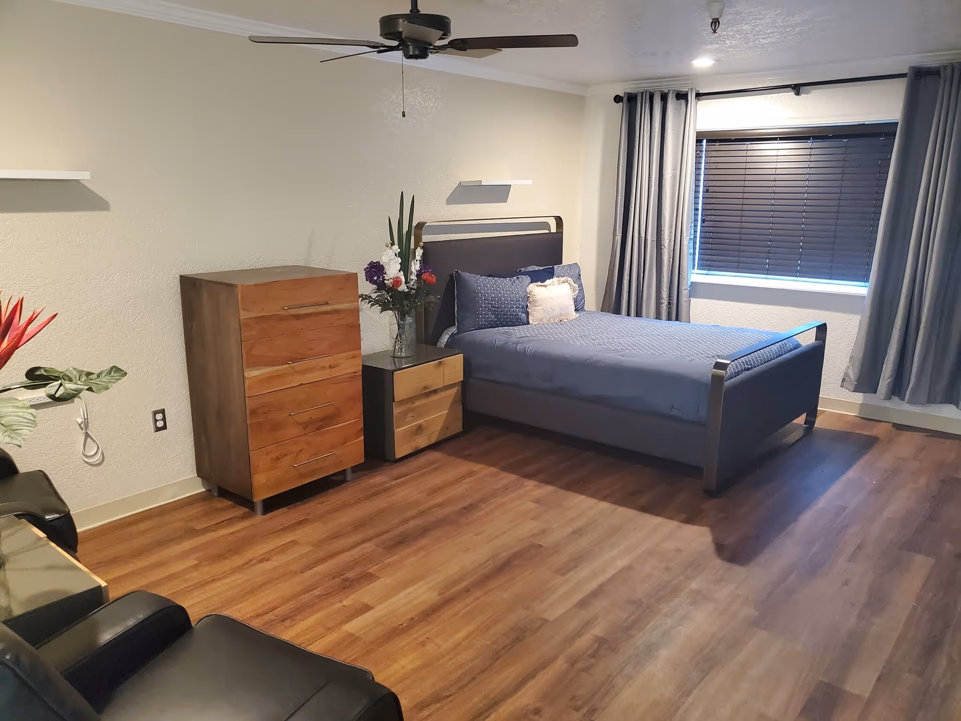 A bedroom with a single bed covered in blue bedding and pillows, a wooden dresser and nightstand with a vase of flowers, a ceiling fan, a window with closed blinds and gray curtains, and two black chairs partially visible in the foreground. The room has wood flooring and light-colored walls.