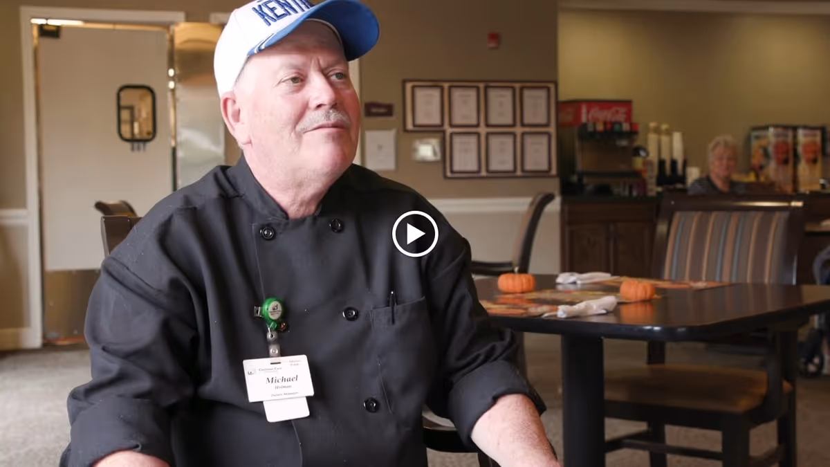 A man wearing a black chef's coat and a white and blue cap sits at a table in a dining area. The man has a name tag that reads 'Michael' and appears to be a kitchen manager. The dining room has tables set with napkins and small decorative pumpkins. In the background, there is a beverage station and another person seated at a table.