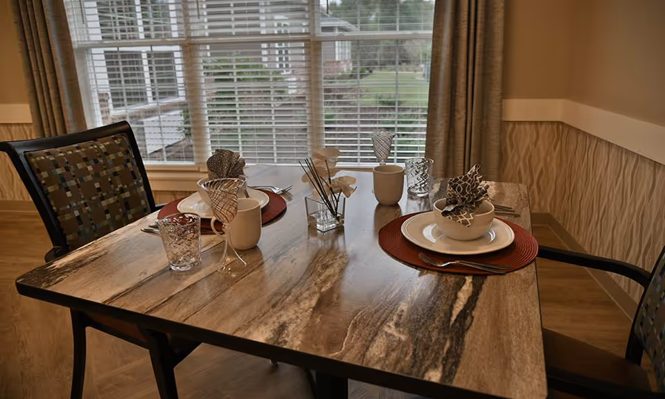 A dining table set for two with patterned napkins, white plates, bowls, cups, and glasses on red placemats. The table is near a window with blinds and beige curtains, with a small decorative vase with flowers in the center.
