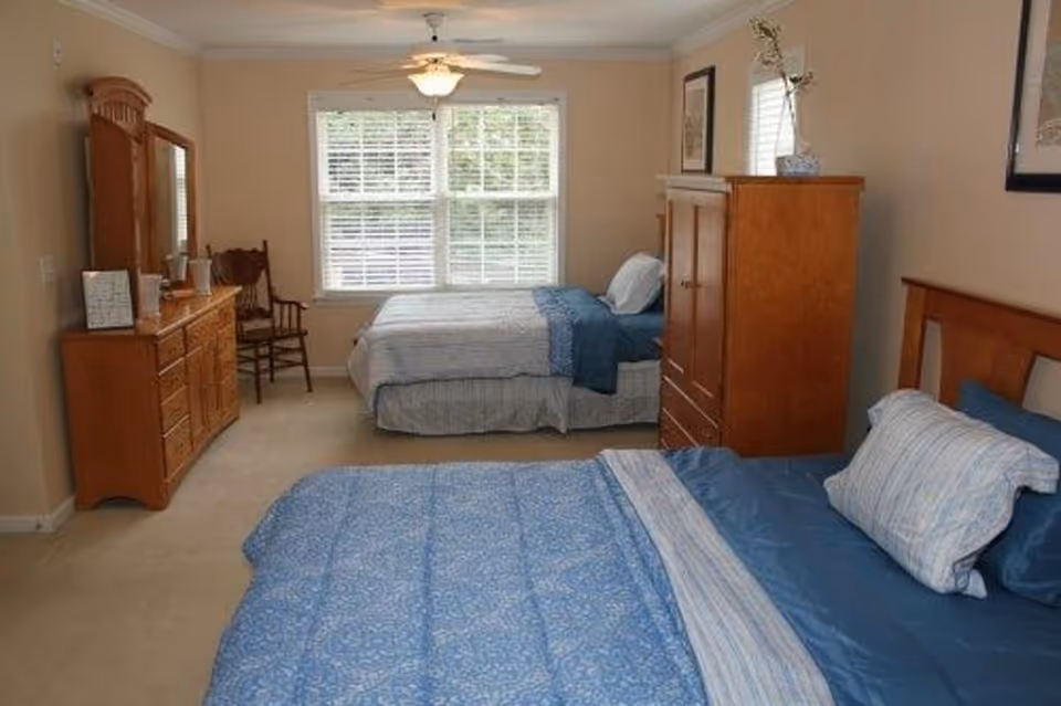 A bedroom with two beds, each made with blue and white bedding. There is a wooden dresser with a mirror on the left side of the room, a wooden chair next to it, and a wooden armoire between the beds. A large window with white blinds is on the far wall, letting in natural light.
