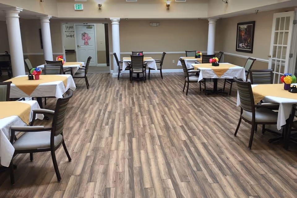 A dining room with several tables covered with white and yellow tablecloths, each table decorated with colorful flower centerpieces. The room has wooden flooring, beige walls, white columns, and framed artwork on the walls. There are multiple chairs around each table and an exit sign above a door at the back.
