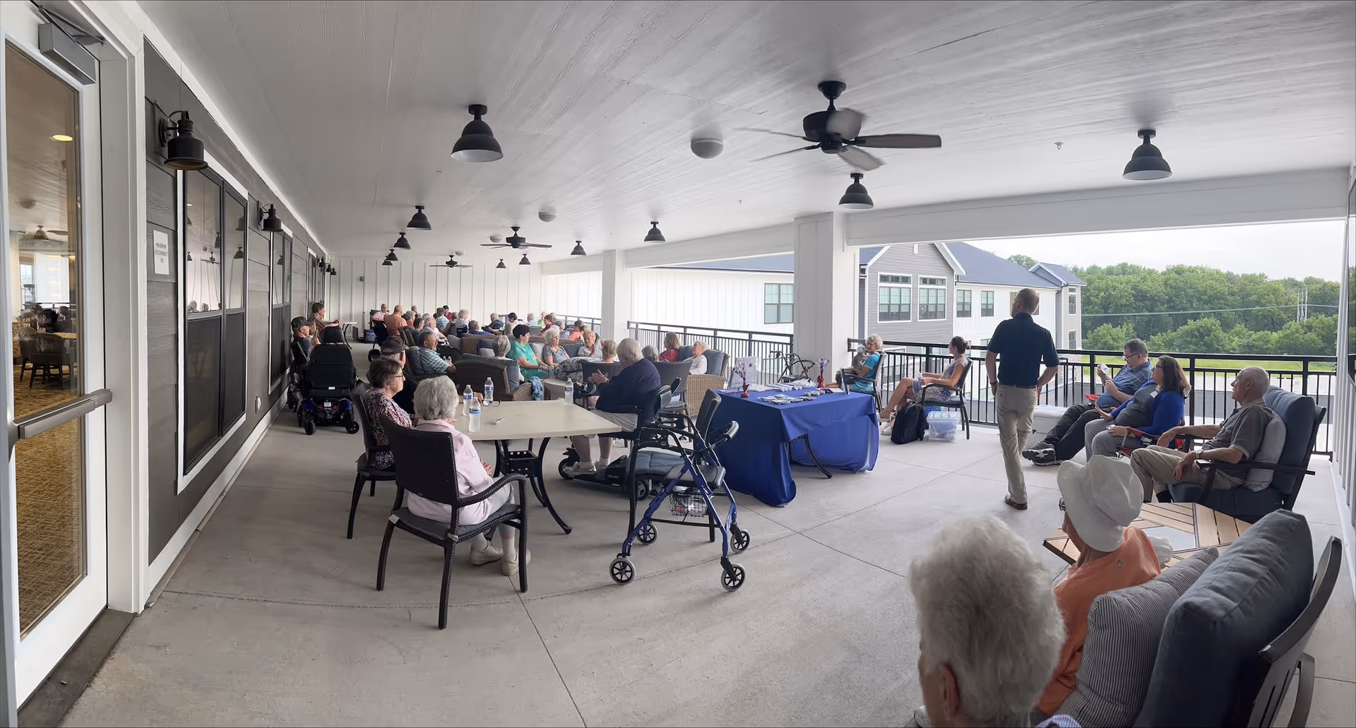 A covered outdoor patio area at a senior living facility with many elderly residents seated at tables and chairs, some using walkers and wheelchairs. The patio overlooks a green landscape and nearby buildings. Ceiling fans and lights are mounted on the white ceiling, and a table with a blue tablecloth is set up near the railing.