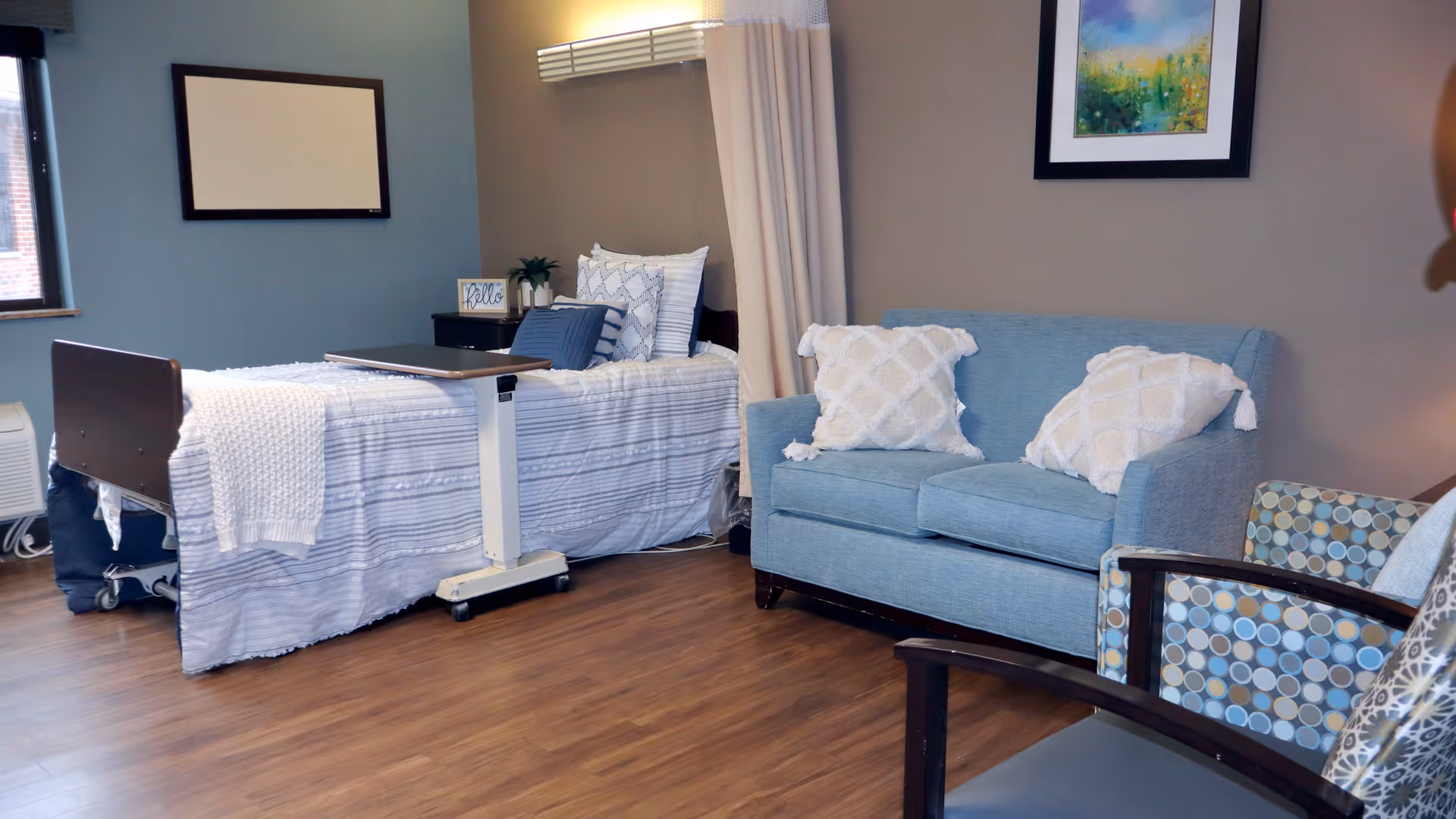 A furnished senior bedroom with a hospital-style bed, overbed table, blue loveseat and chairs on wood flooring.