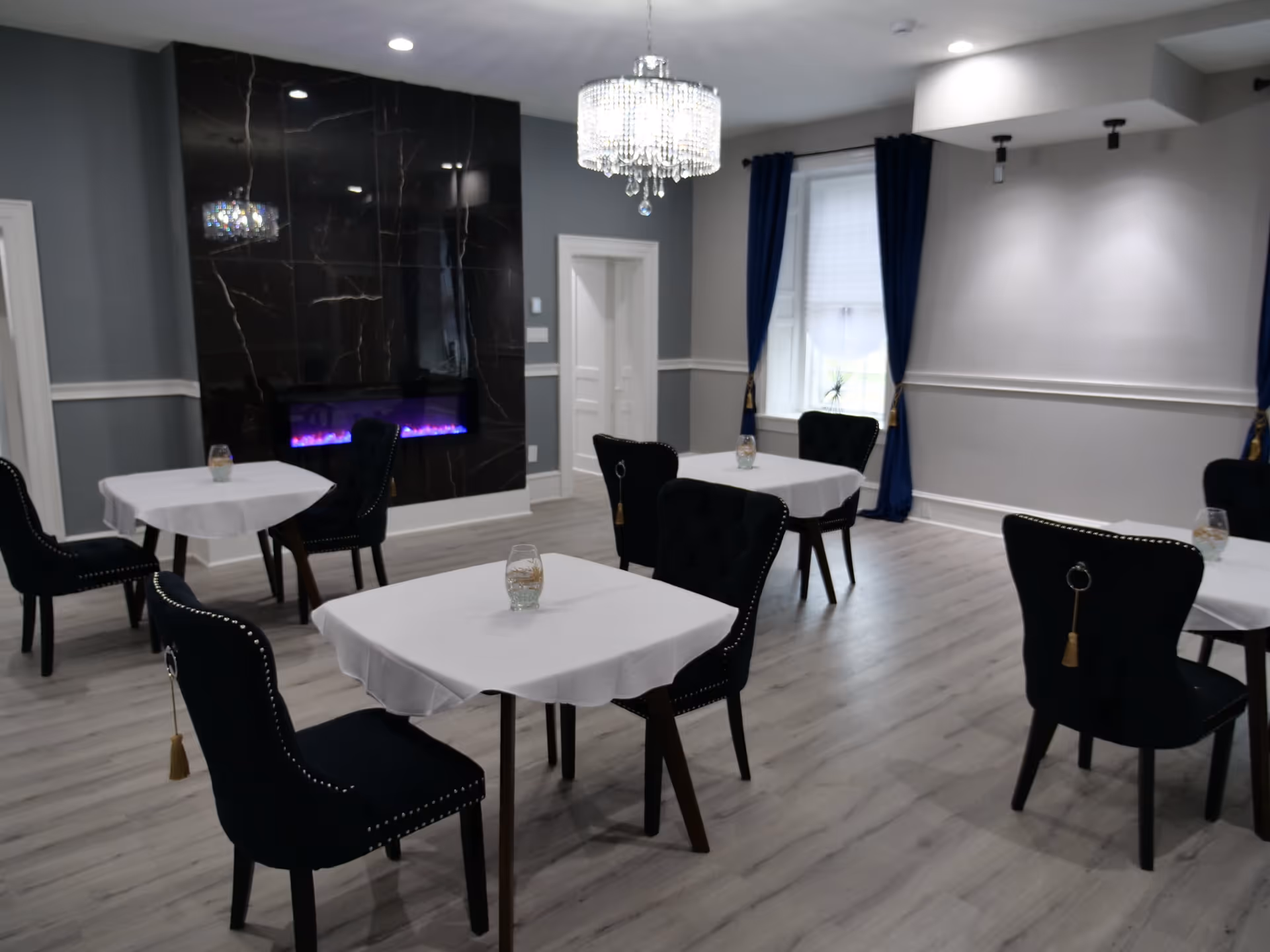 A dining room with several small square tables covered with white tablecloths, each accompanied by black upholstered chairs with decorative tassels. The room features light wood flooring, gray walls with white trim, a large window with blue curtains, a modern black marble fireplace with a purple flame, and a crystal chandelier hanging from the ceiling.