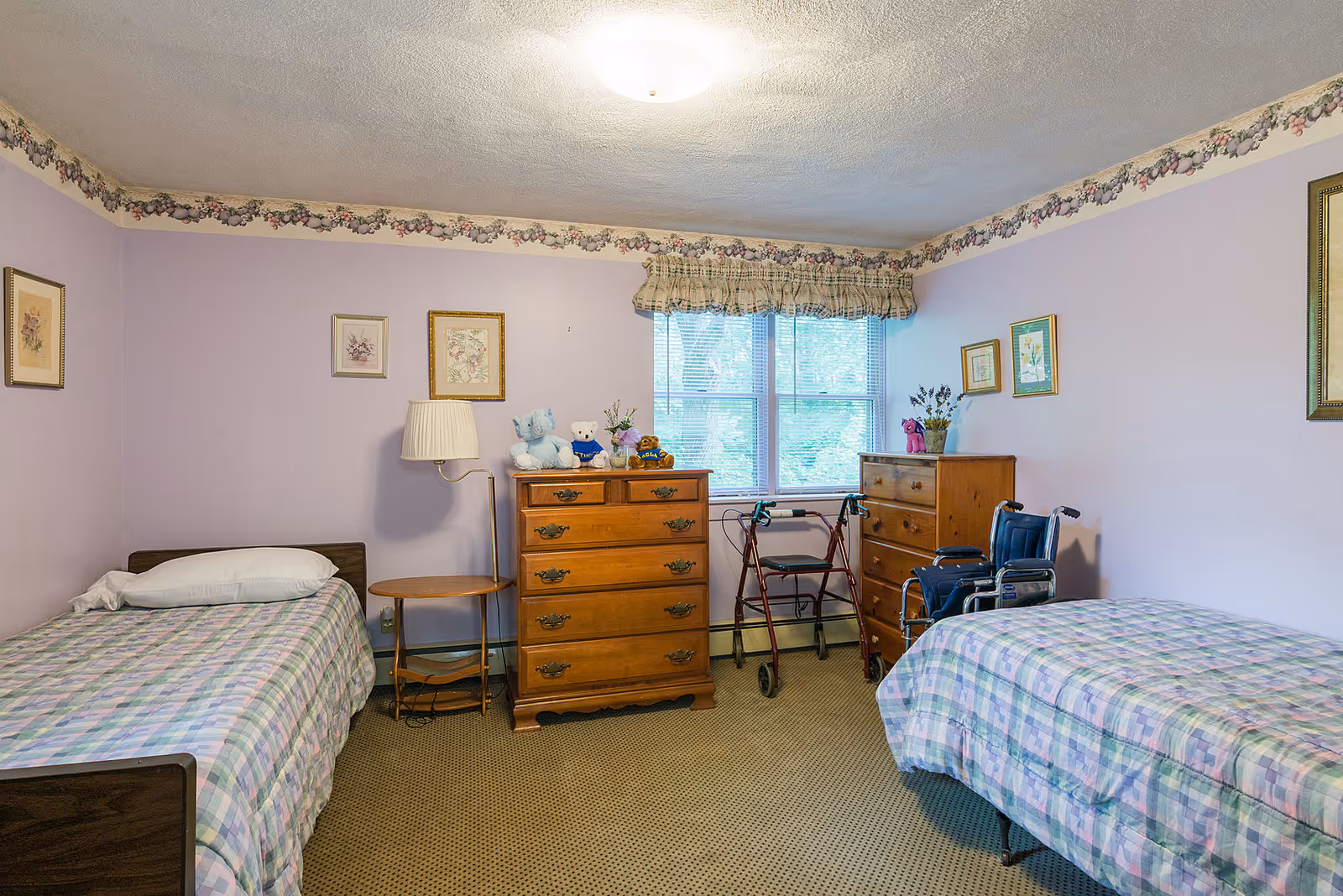 A pastel purple bedroom with two twin beds, wooden dressers, and mobility aids (walker and wheelchair) under a window.