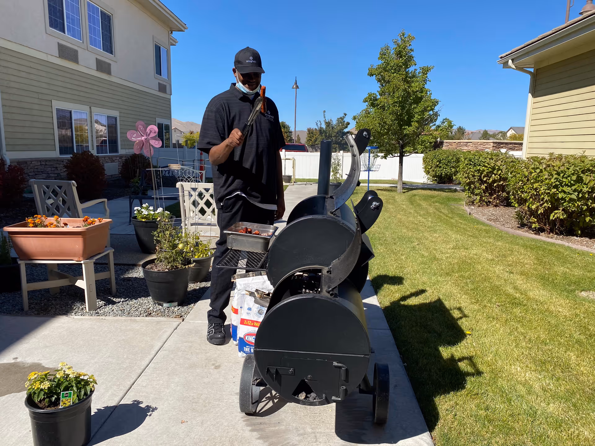 A man wearing a black shirt, black pants, and a black cap is standing outdoors next to a large black barbecue smoker grill. He is holding a pair of tongs with a sausage on them. The setting is a sunny day with a clear blue sky, green grass, potted plants, and residential-style buildings in the background.