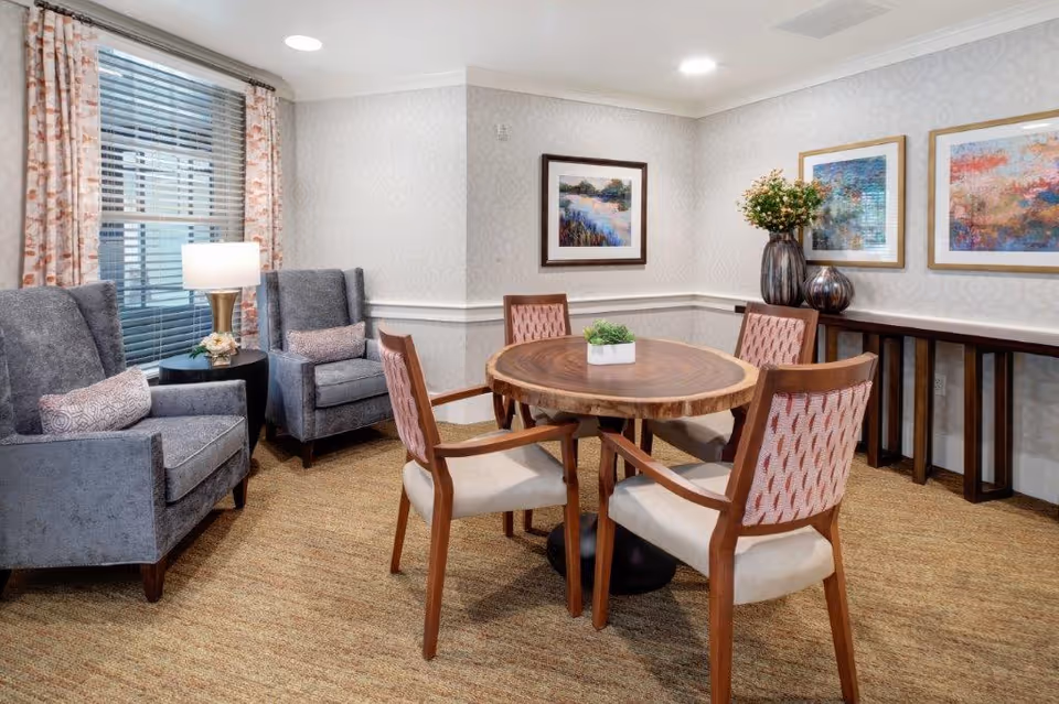 A cozy sitting area with a round wooden table surrounded by four chairs with patterned backs. To the left, there are two gray upholstered armchairs with small pillows, next to a side table with a lamp and a small flower arrangement. The walls are decorated with three framed paintings and a large vase with flowers on a narrow console table. The room has carpeted flooring and a window with blinds and patterned curtains.