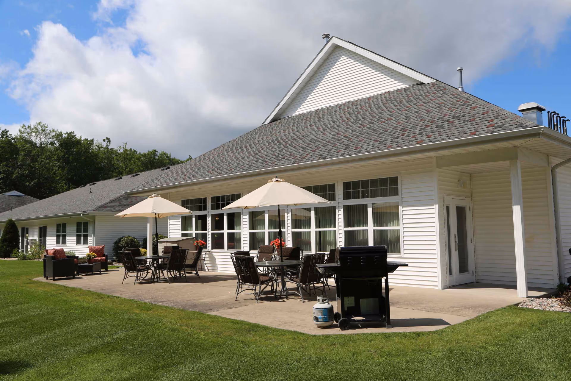 Outdoor patio area of a senior living facility with white siding building in the background, featuring tables with chairs and umbrellas, a grill, and green lawn under a partly cloudy sky.