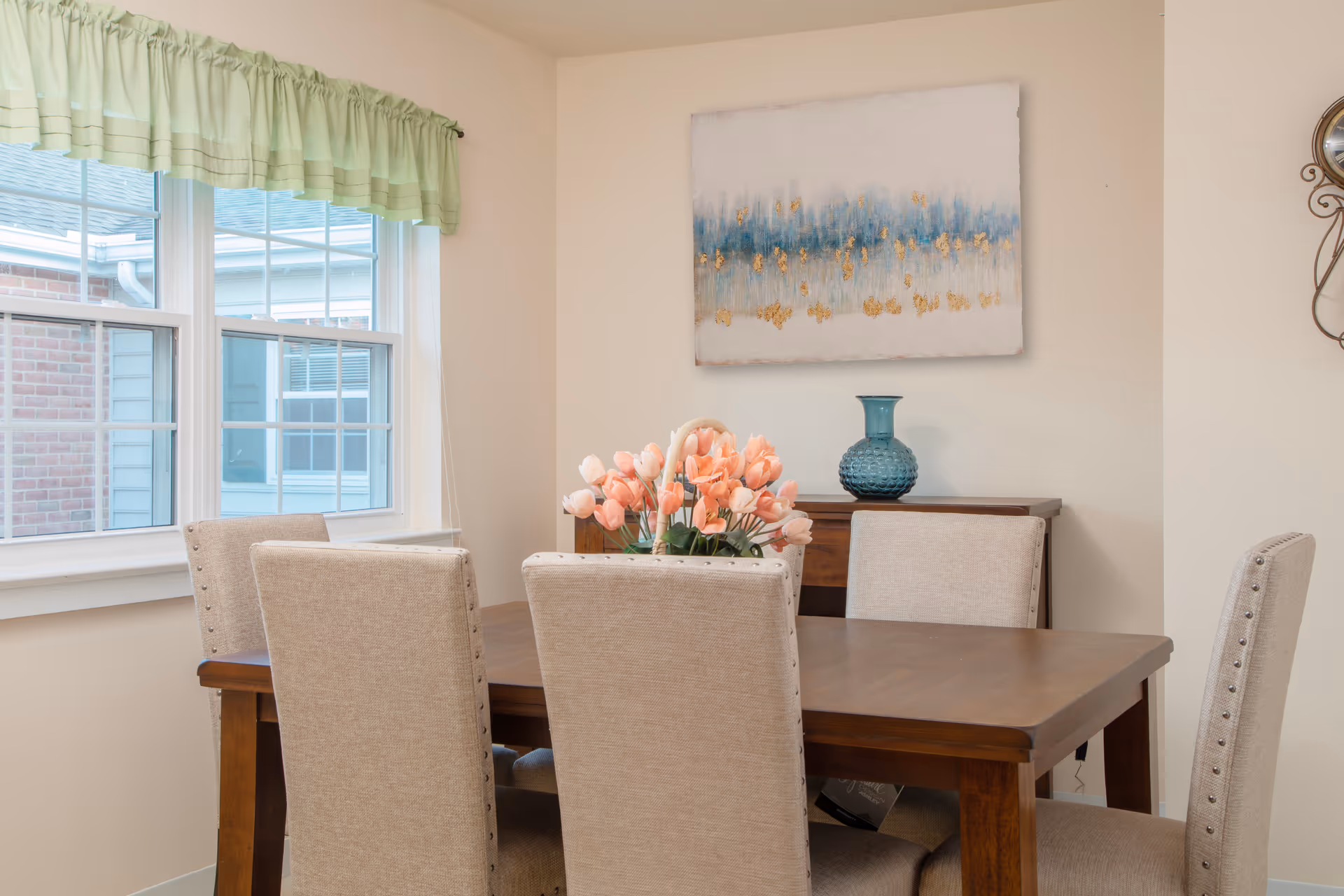 A dining room with a wooden table surrounded by six beige upholstered chairs. On the table is a basket filled with pink tulips. Behind the table is a sideboard with a blue vase and a large abstract painting with blue and gold accents hanging on the wall. A window with a light green valance lets in natural light.