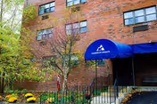 Brick building entrance with blue awning, trees, and a small garden.