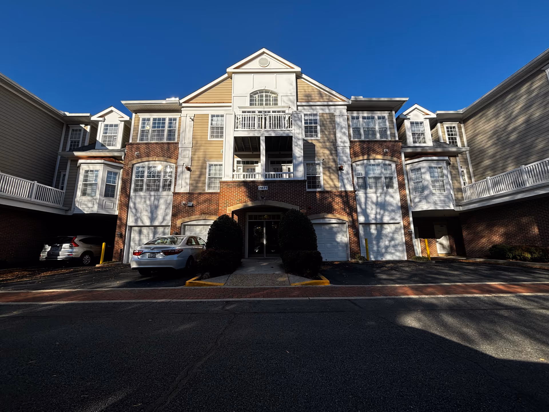 Front exterior view of a multi-story residential building with brick and beige siding, multiple windows, balconies, and a central entrance. Two cars are parked in front of the building under a clear blue sky.