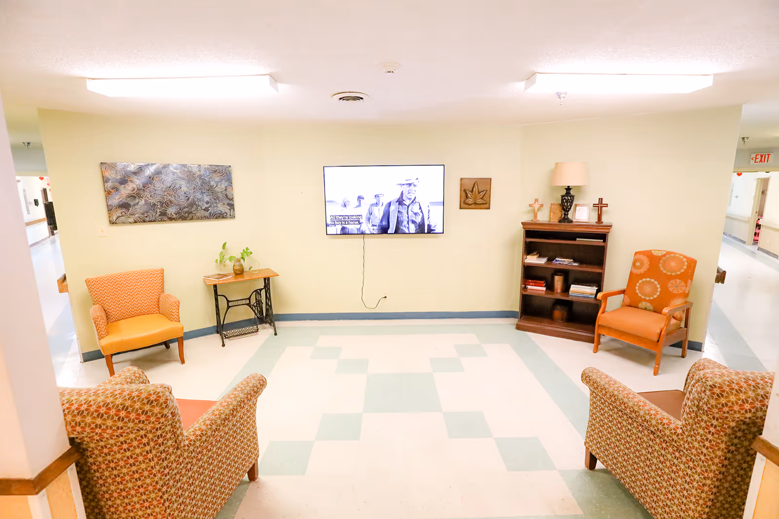 Small communal lounge with four upholstered chairs facing a wall-mounted TV, a bookshelf, and side tables in a brightly lit hallway.