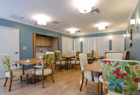 A bright dining room with multiple wooden tables and chairs featuring floral upholstery. The room has light blue walls, wooden flooring, ceiling lights, and large windows with vertical blinds.