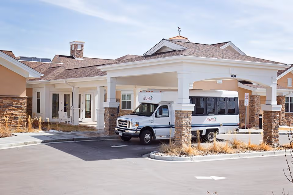 Exterior view of AltaVita Assisted Living facility showing the entrance with a covered drop-off area supported by stone pillars. A white shuttle bus with AltaVita branding is parked under the covered area. The building features beige walls with stone accents and a clear sky above.