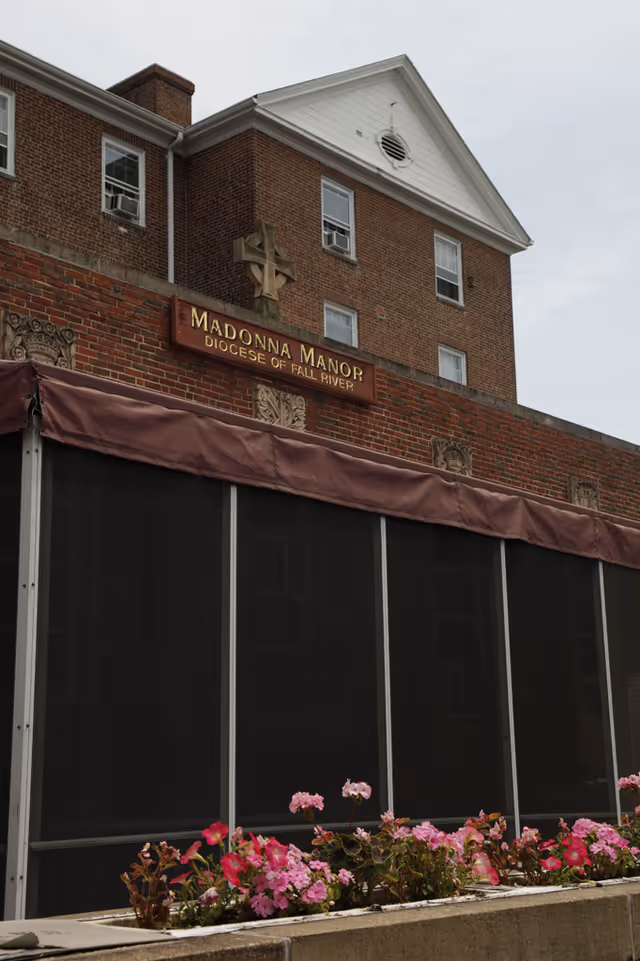 Exterior view of Madonna Manor building showing a brick facade with a sign that reads 'Madonna Manor Diocese of Fall River'. The building has several windows and a white triangular roof section. In front of the building, there is a screened porch area with a maroon awning and a flower bed with pink and red flowers.