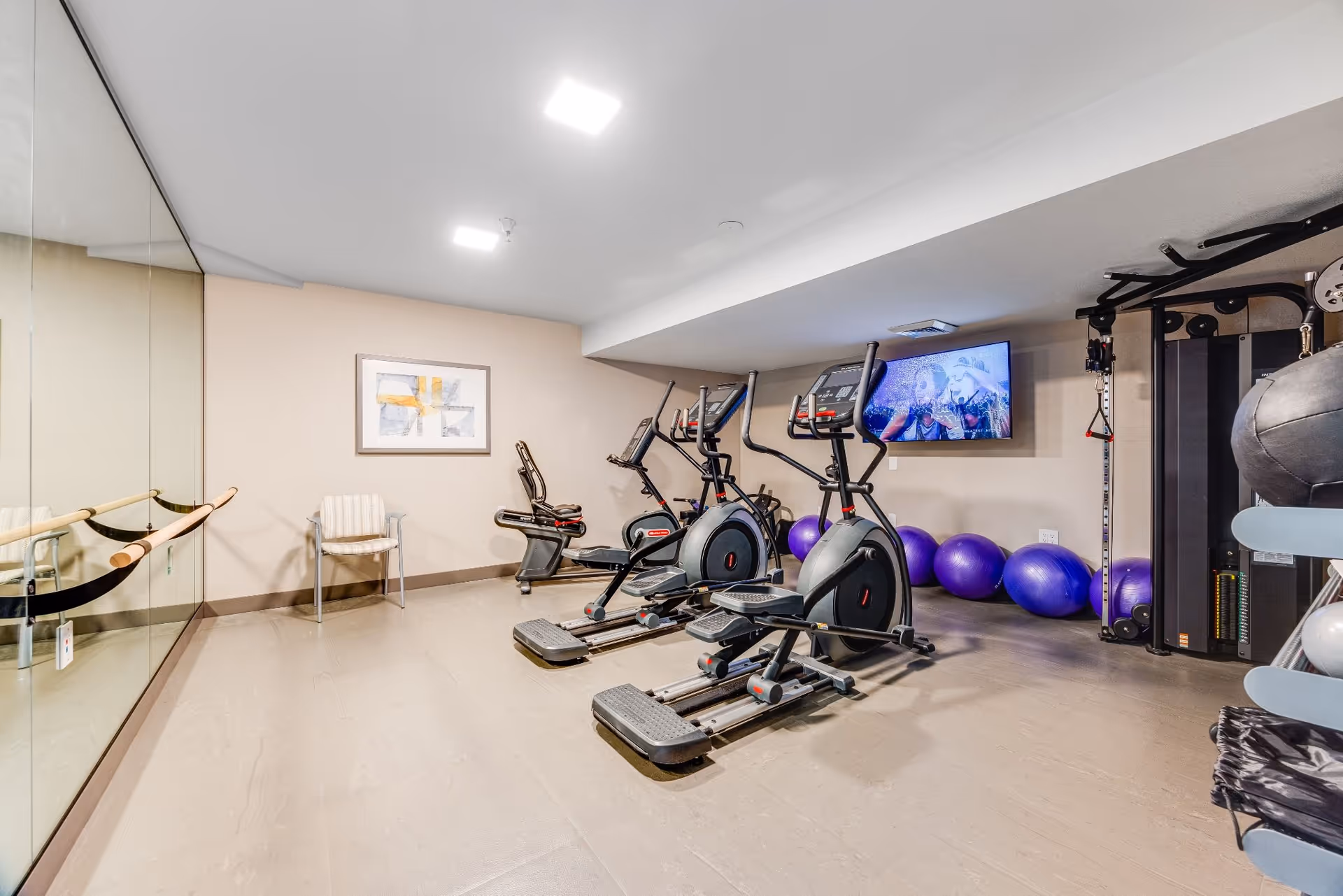 A fitness room with exercise equipment including elliptical machines, a recumbent bike, purple exercise balls, a multi-function weight machine, and a wall-mounted TV. There is a large mirror on one wall with a ballet barre, a chair, and a framed abstract artwork on the opposite wall.