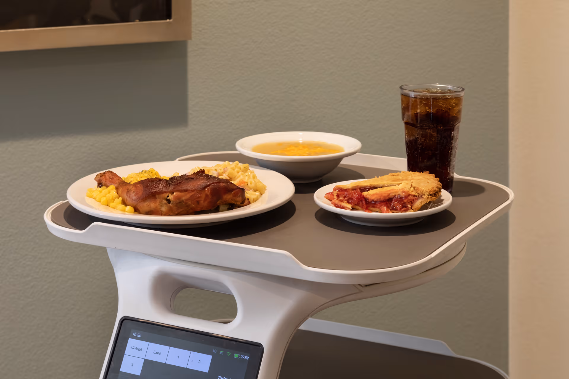 A meal on a tray with a plate of roasted chicken, corn, and macaroni and cheese, a bowl of soup, a slice of cherry pie on a small plate, and a glass of soda, set against a plain wall background.