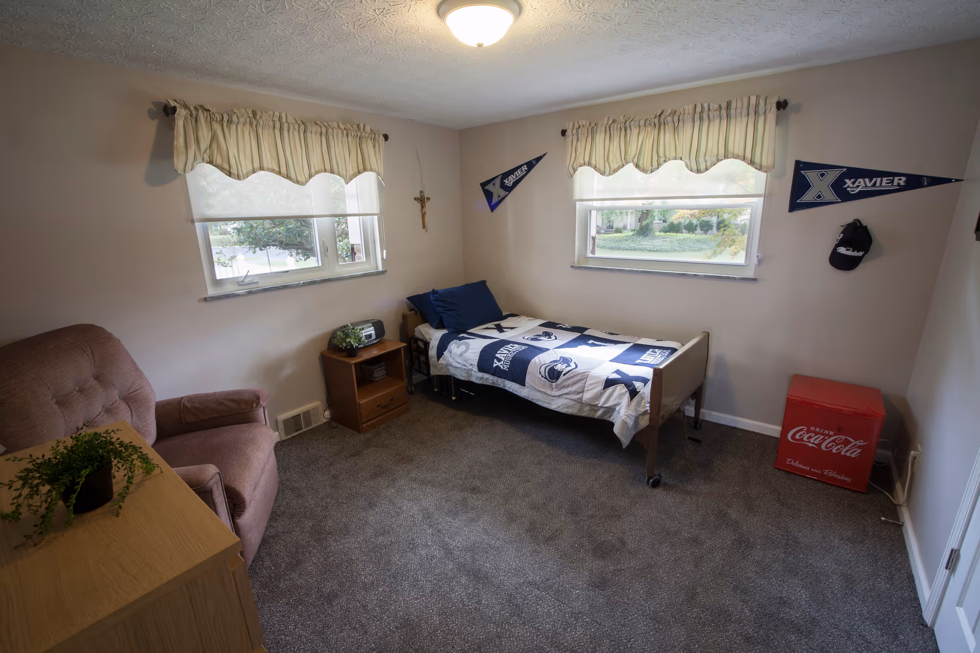A small bedroom with a single bed covered in a blue and white Xavier University themed blanket and pillows. The room has two windows with striped valances, a brown armchair, a wooden nightstand with a radio and a small plant, a red Coca-Cola mini fridge, and two Xavier pennants on the walls. The carpet is gray and the walls are light beige.