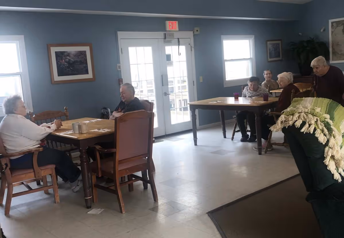 A common area in Margaret's Meadows with elderly residents sitting at two wooden tables. The room has blue walls, two windows, a set of glass double doors with an exit sign above, and framed pictures on the walls. There is a green armchair with a green and white throw blanket in the foreground.