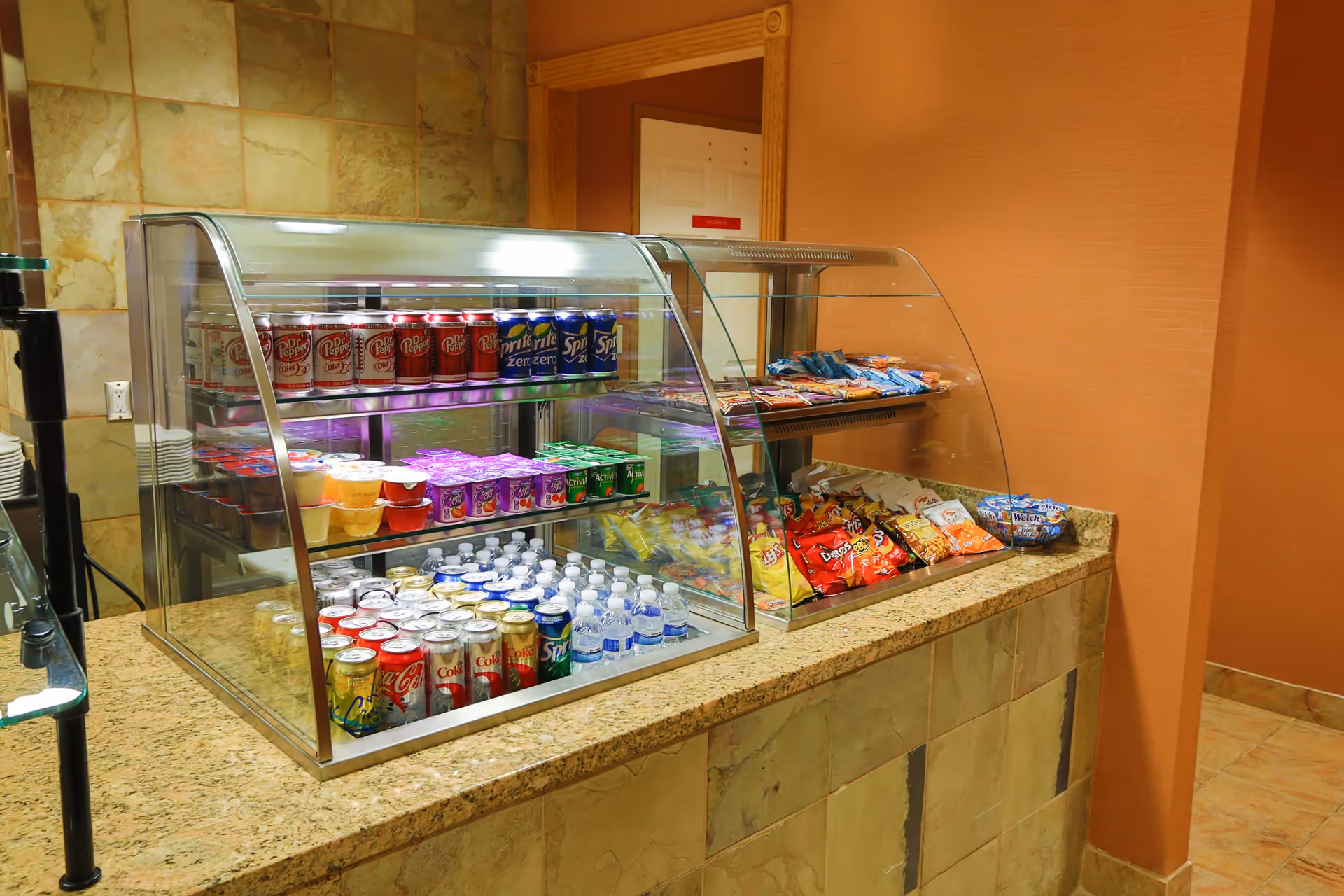 A snack and beverage counter with two glass display cases. One case contains canned sodas, bottled water, and small cups of yogurt and fruit. The other case holds various bags of chips and snacks. The counter has a beige tiled front and a granite countertop, with a warm orange wall in the background.