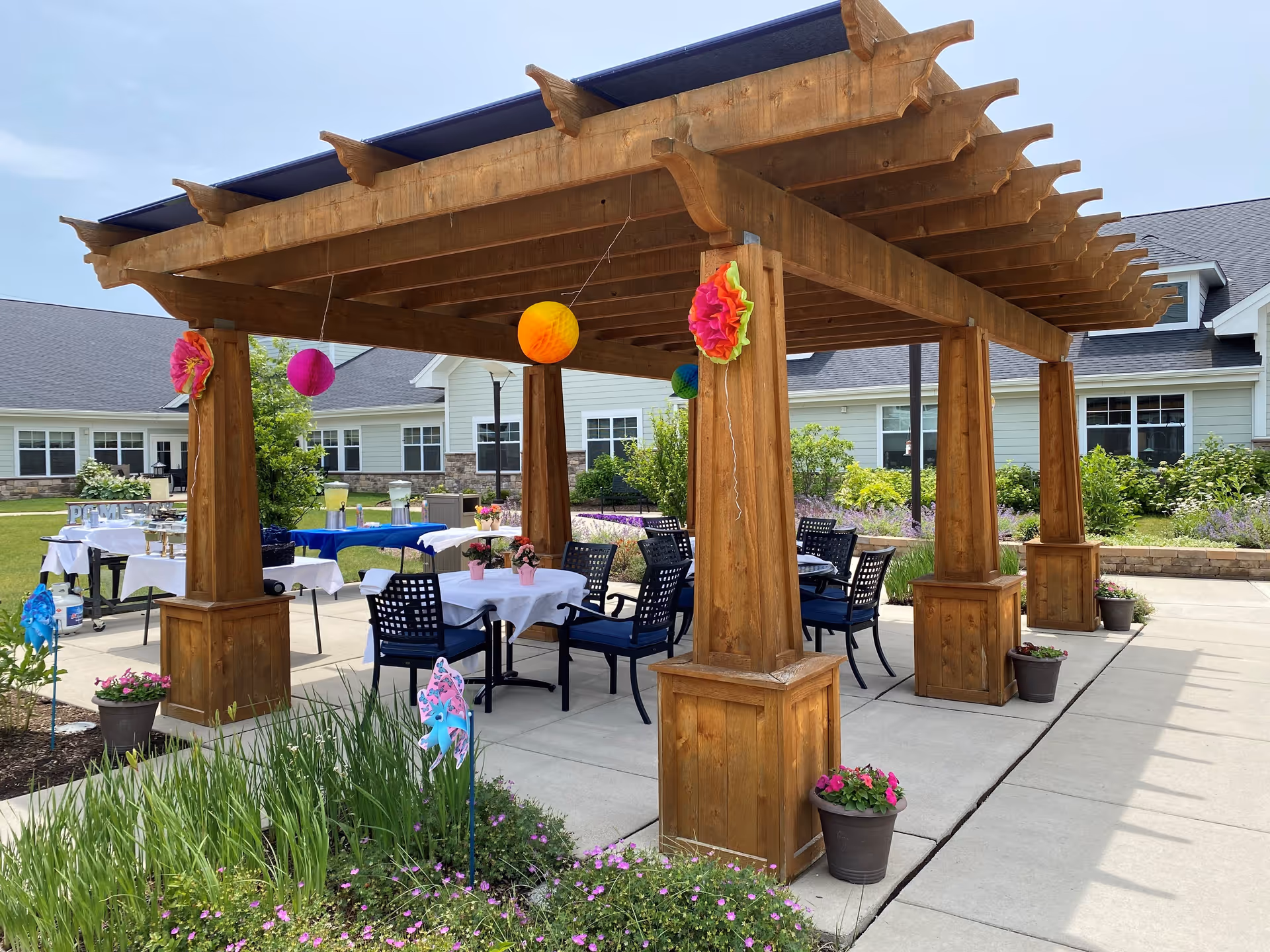 Outdoor patio area at Pomeroy Senior Living of Northville with a wooden pergola decorated with colorful hanging paper lanterns and flowers. Under the pergola are tables with white tablecloths and chairs, surrounded by flower pots and greenery. In the background, there are buildings with light green siding and multiple windows.