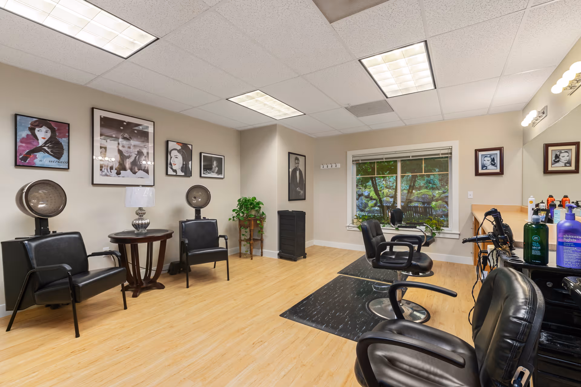 Interior of a salon room with black salon chairs, hair dryers, and a large window showing greenery outside. The room has light wood flooring, beige walls decorated with framed portraits, and a small table with a lamp between two chairs.