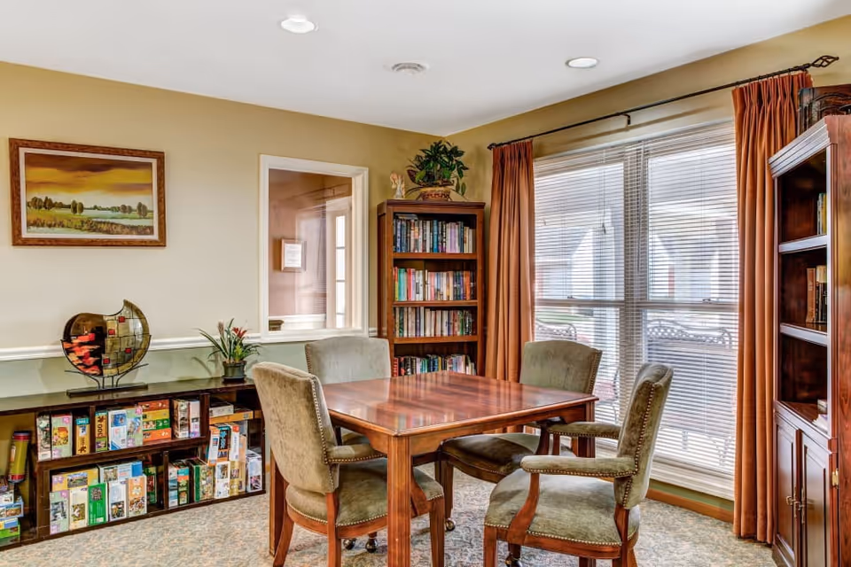 A cozy room with a wooden table surrounded by four upholstered chairs. There are two bookshelves filled with books and board games, a large window with blinds and rust-colored curtains, and a framed landscape painting on the wall.