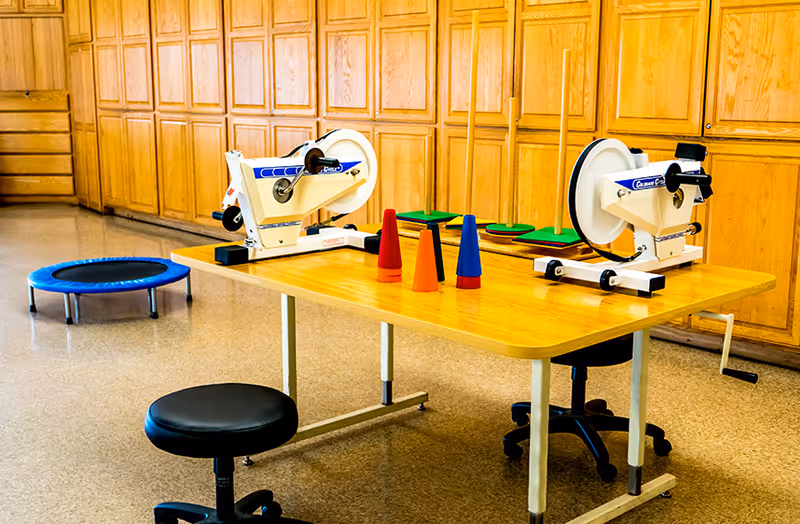 A therapy room with a wooden table holding two hand-crank exercise machines and several colorful cones. There are two black stools around the table and a small blue trampoline on the floor. The background features wooden cabinets covering the wall.