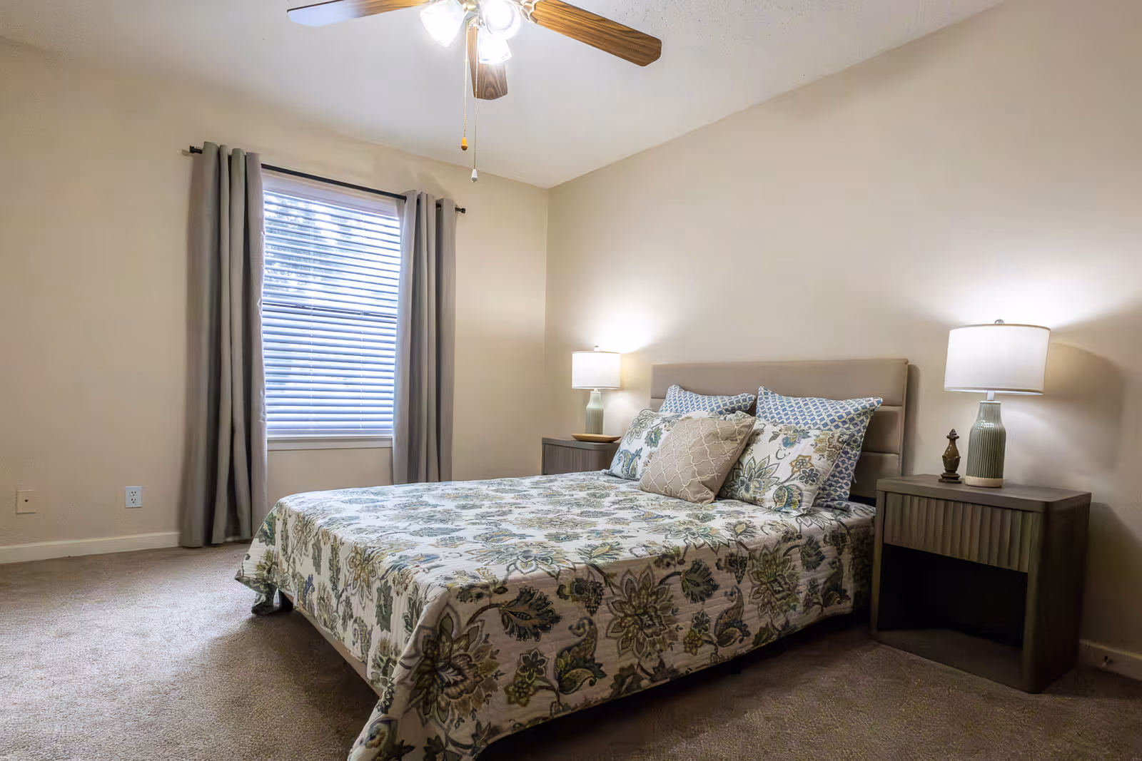 A bedroom with a bed covered in a floral patterned bedspread and multiple pillows. There are two nightstands on either side of the bed, each with a lamp. A window with closed blinds and gray curtains is on the left wall. The room has beige walls and carpeted floor, with a ceiling fan overhead.