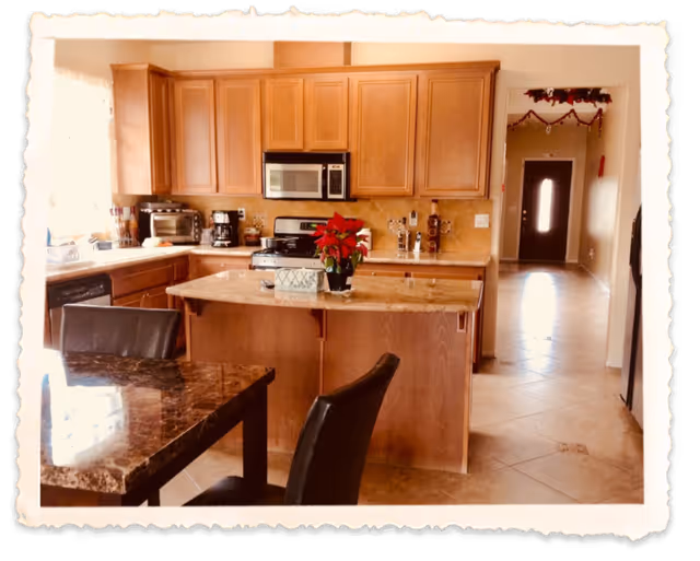 A warm-toned kitchen with wooden cabinets, a marble countertop island with a red poinsettia plant, and a dining table with black chairs. The kitchen includes a microwave, stove, coffee maker, and toaster oven. A hallway with tile flooring and a door at the end is visible in the background, decorated with holiday garlands.