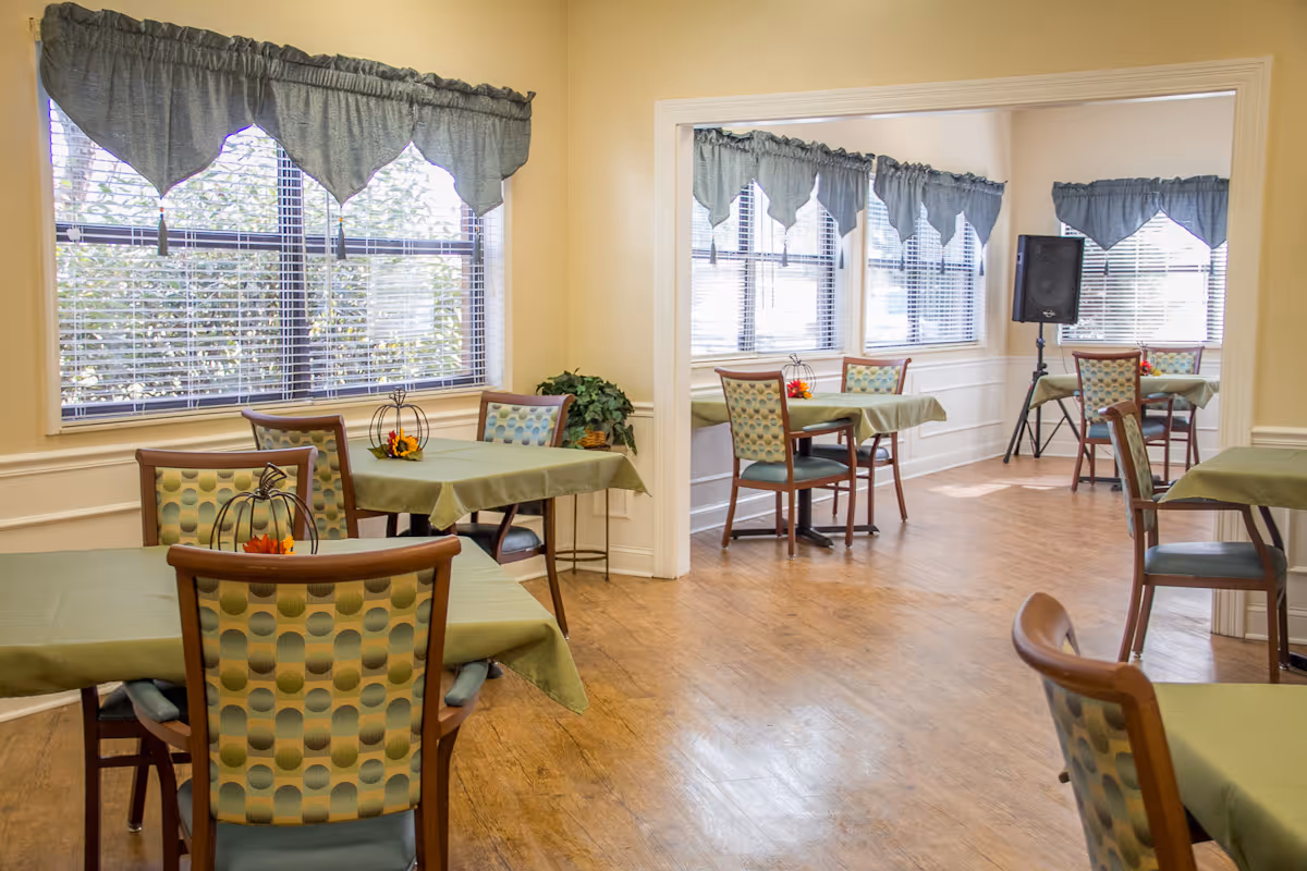 A bright dining area with multiple tables covered in green tablecloths and chairs with patterned upholstery. Large windows with blue valance curtains let in natural light, and there are small decorative centerpieces on the tables. The room has wood flooring and light-colored walls.