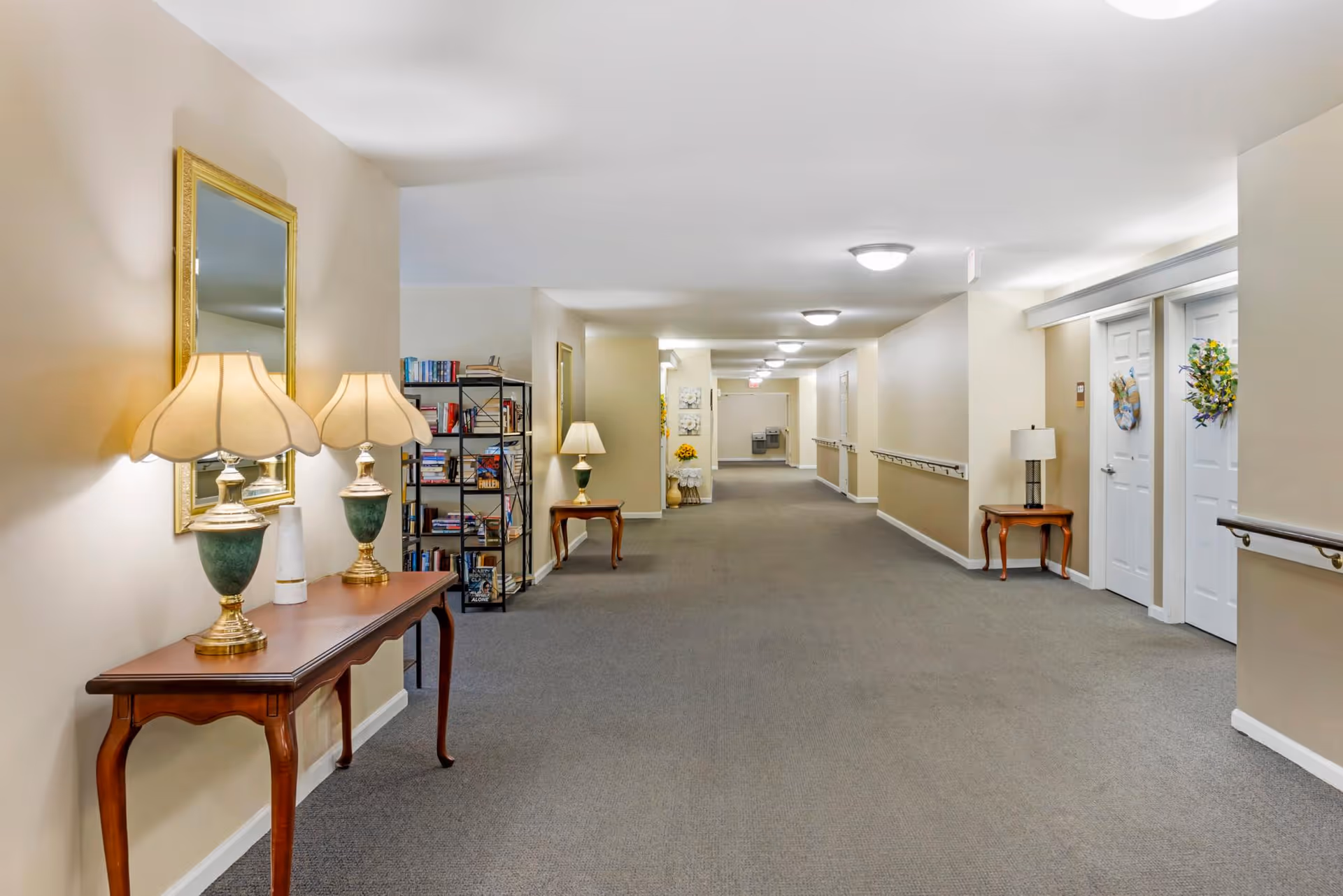 A long, well-lit hallway in a senior living facility with beige walls and carpeted floor. The hallway features wooden tables with lamps and decorative items, a bookshelf filled with books, handrails along the walls, and doors decorated with wreaths. Ceiling lights illuminate the corridor evenly.