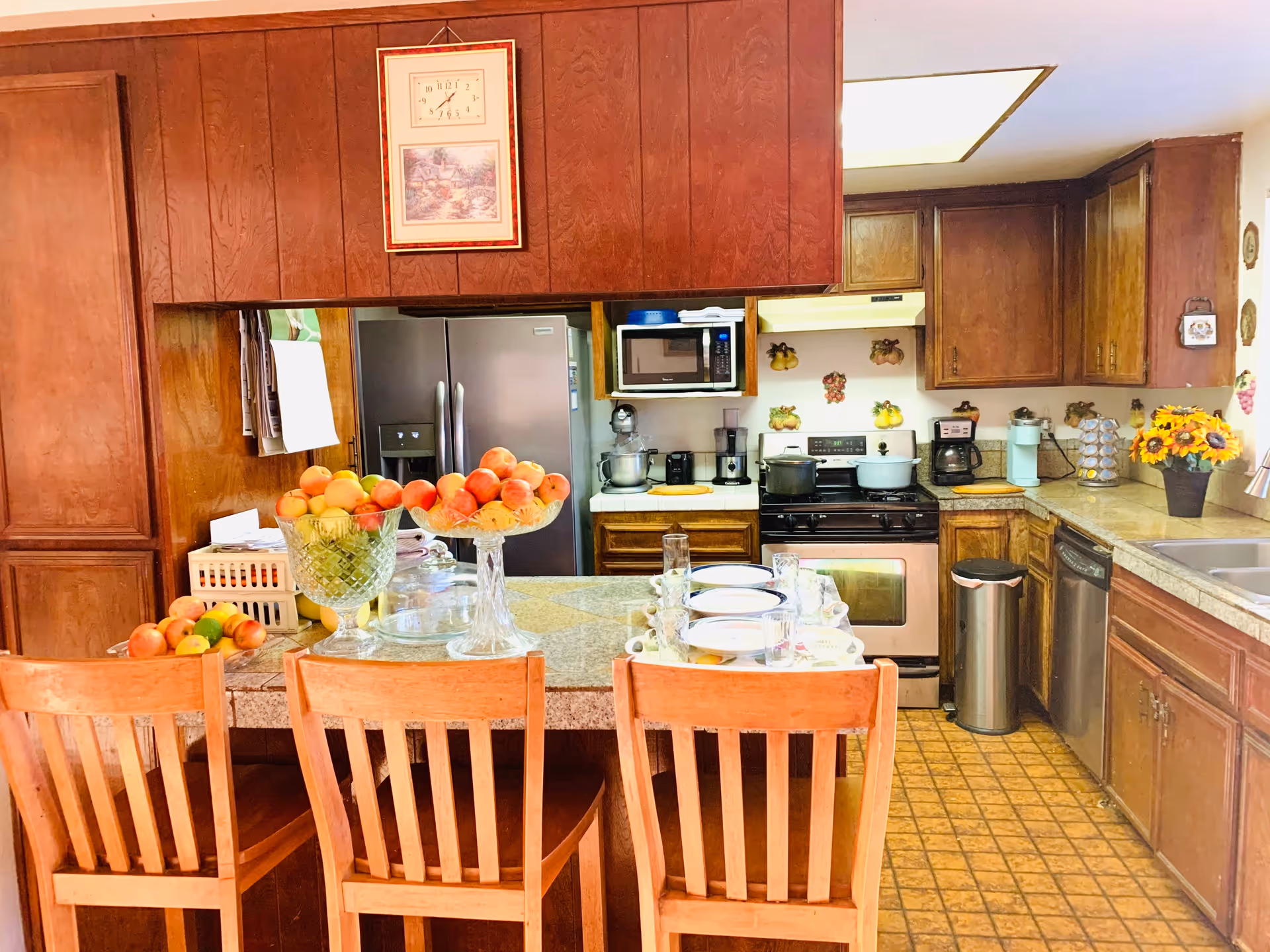 A kitchen with wooden cabinets and a tiled floor. There is a kitchen island with three wooden chairs, topped with bowls of fruit and place settings with plates, glasses, and napkins. The kitchen has a stainless steel refrigerator, microwave, stove with pots, coffee maker, and a dishwasher. A clock and framed picture hang on the wooden panel wall above the island.