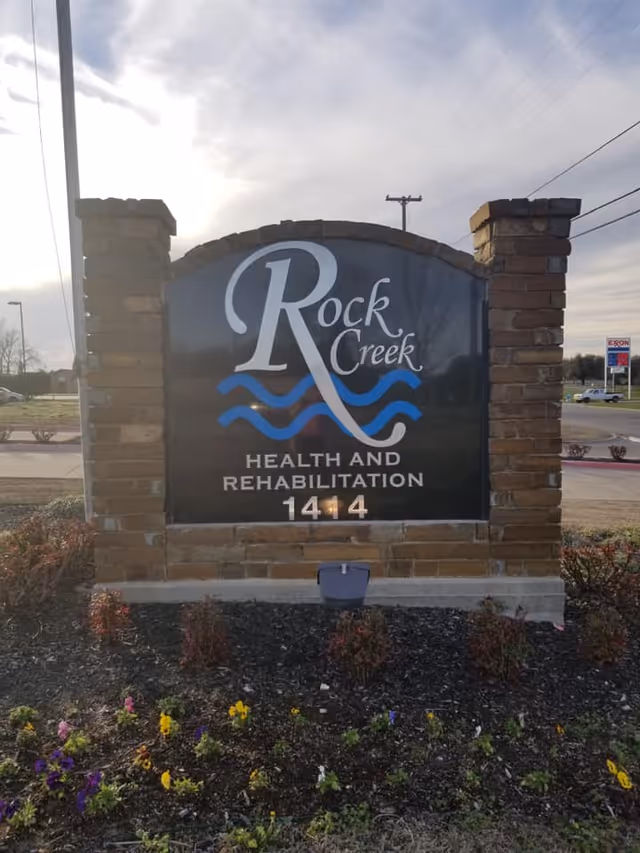 A brick and stone sign for Rock Creek Health and Rehabilitation with the address number 1414. The sign features a large stylized 'R' and blue wave graphics. There are flowers planted in front of the sign and a road with cars and an Exxon gas station in the background under a cloudy sky.