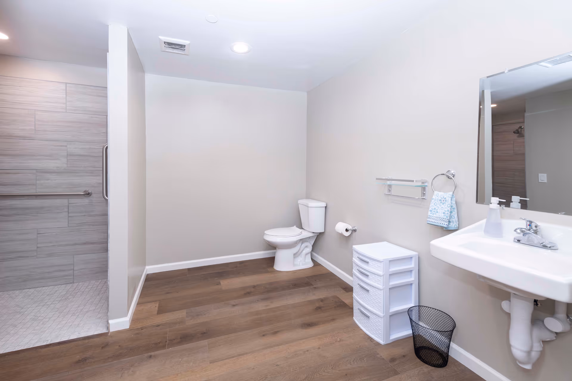 A clean and spacious bathroom with light-colored walls and wood-style flooring. The room features a white toilet, a white sink with a mirror above it, a towel ring with a blue patterned hand towel, a small white storage drawer unit, a black mesh wastebasket, and a walk-in shower with gray tiles and a grab bar.