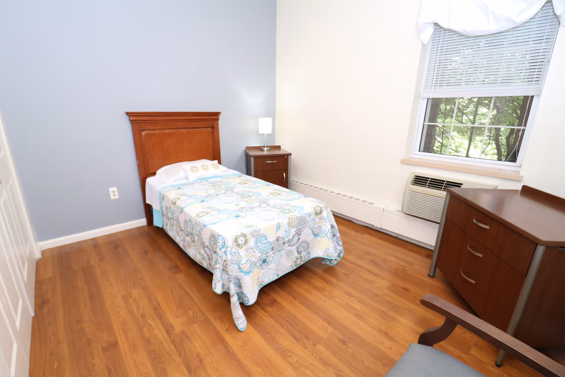 A small bedroom with a single bed, dresser, nightstand, and window.
