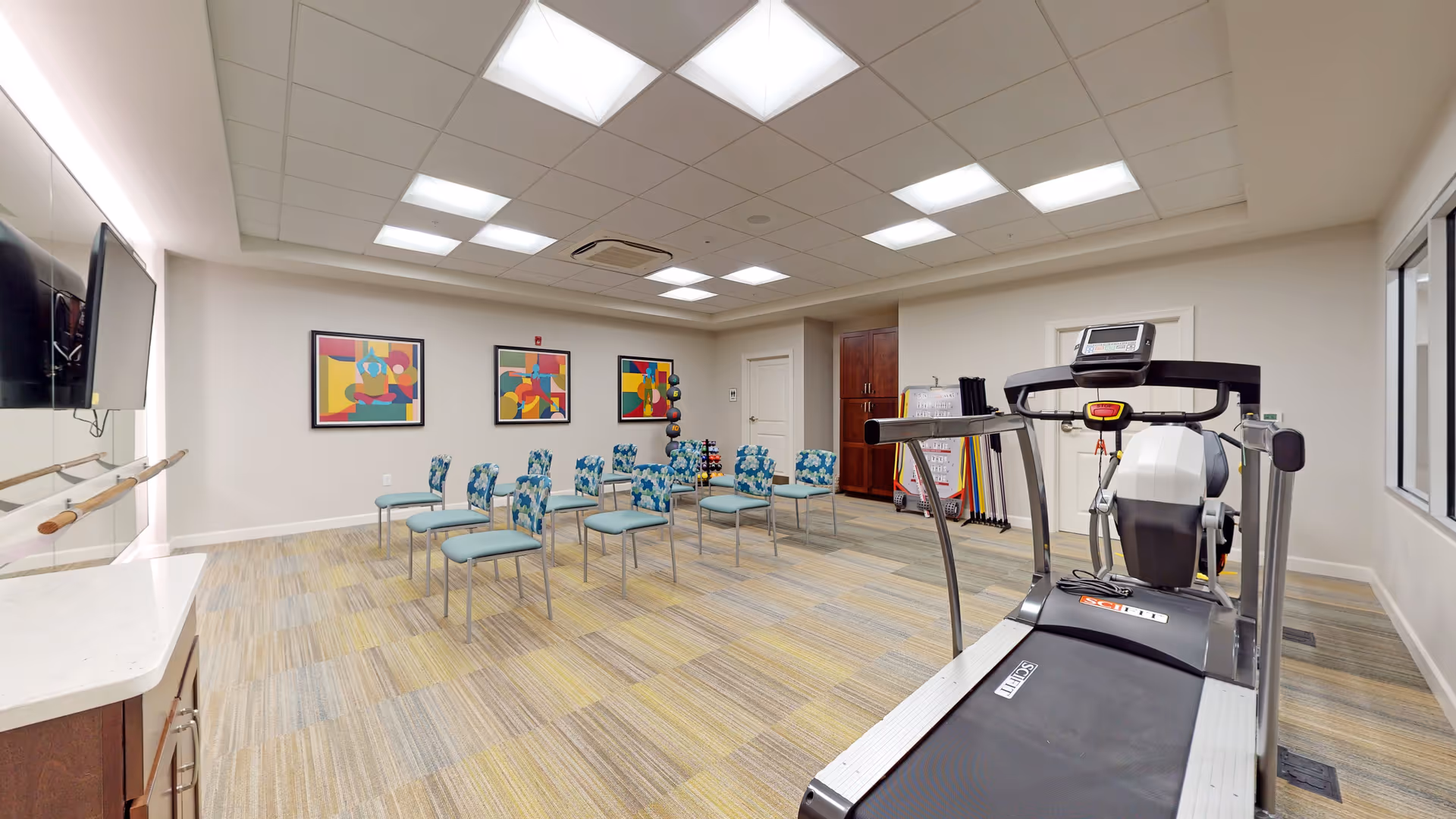 A bright, spacious exercise room with a treadmill in the foreground, several chairs arranged in rows, colorful abstract paintings on the wall, exercise equipment like weights and resistance bands in the corner, and a large mirror with a ballet barre on the left side.