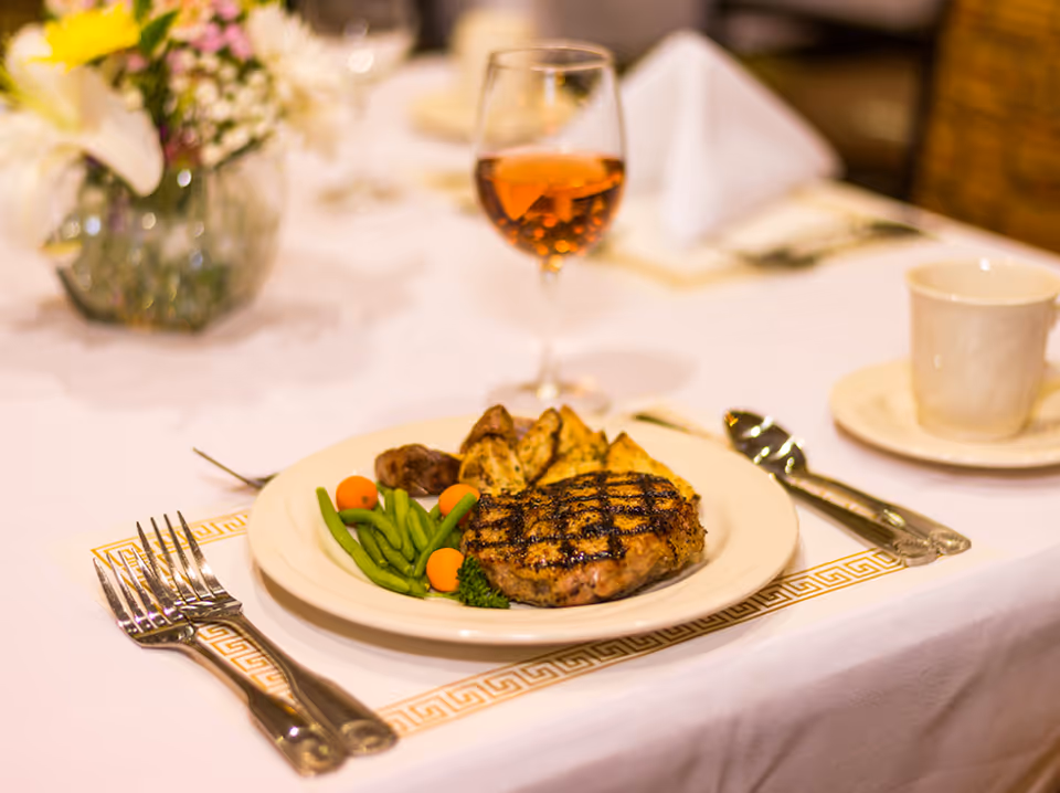A plated meal on a white tablecloth featuring grilled steak, green beans, carrots, and roasted potato wedges, accompanied by a glass of iced tea and a white cup on a saucer, with a floral centerpiece in the background.