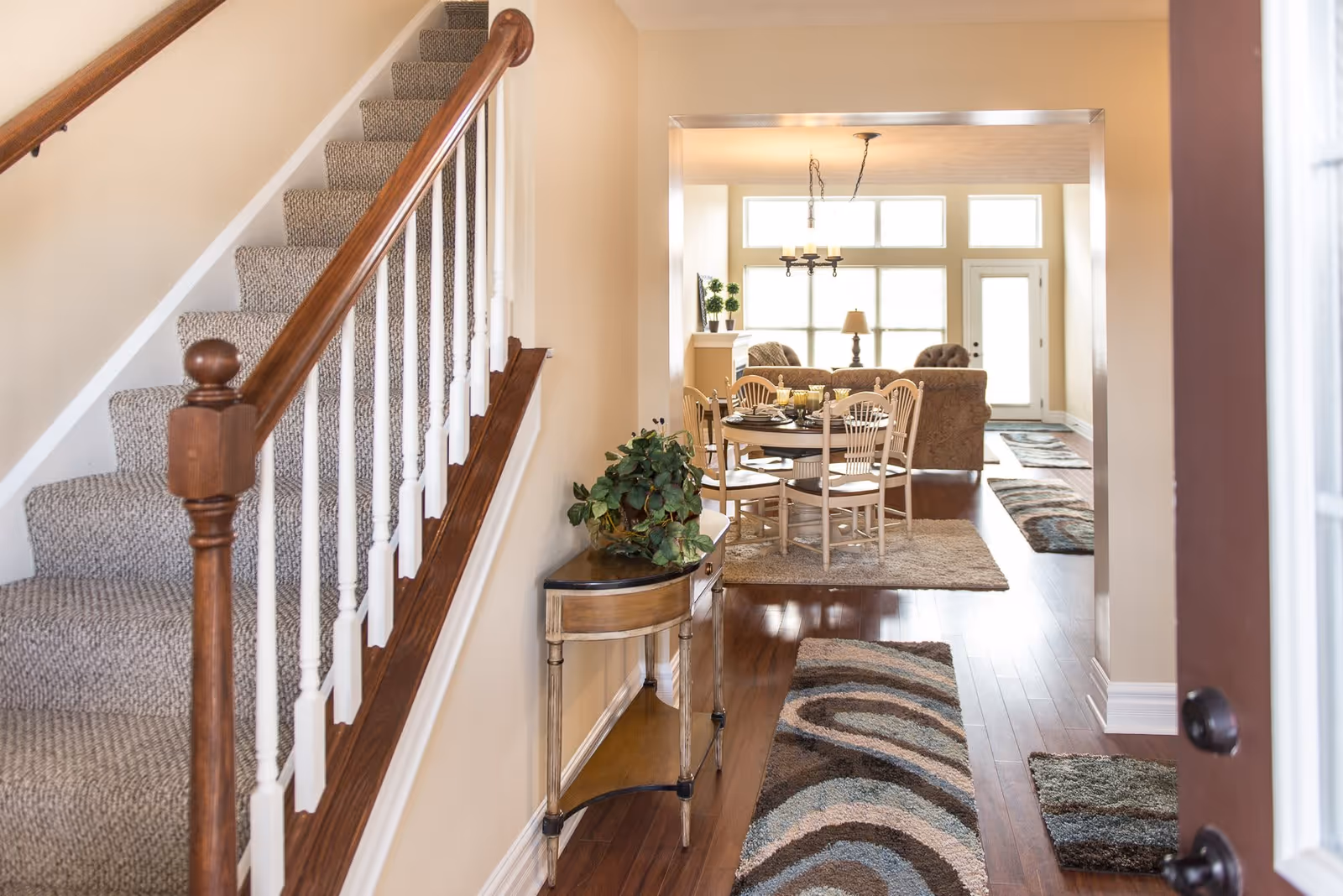 View of a cozy interior space featuring a carpeted staircase with wooden handrails on the left, a small wooden console table with a green plant against the wall, and a dining area with a round table and chairs in the background. The room has hardwood floors with patterned rugs and large windows letting in natural light.